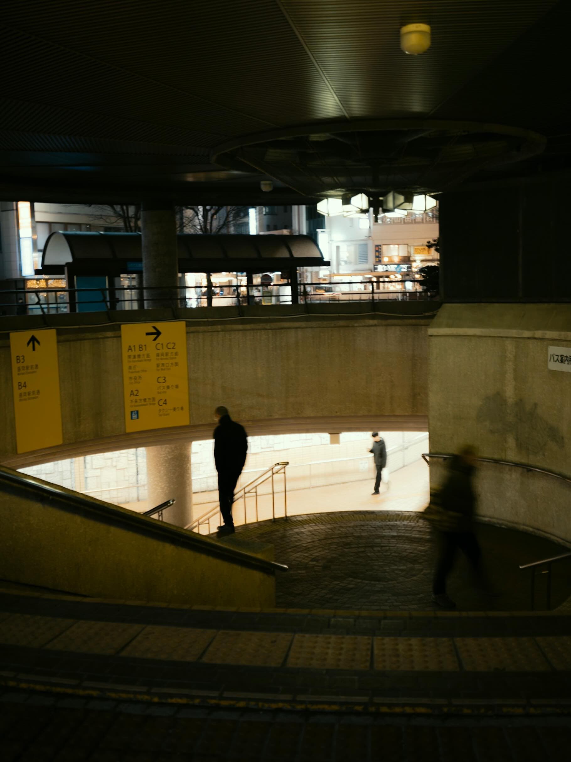 夜の駅前デッキから見下ろした階段と通路。黄色い案内板に「B3」「B4」「A1 B1」などの出口番号が並び、数人が歩いている