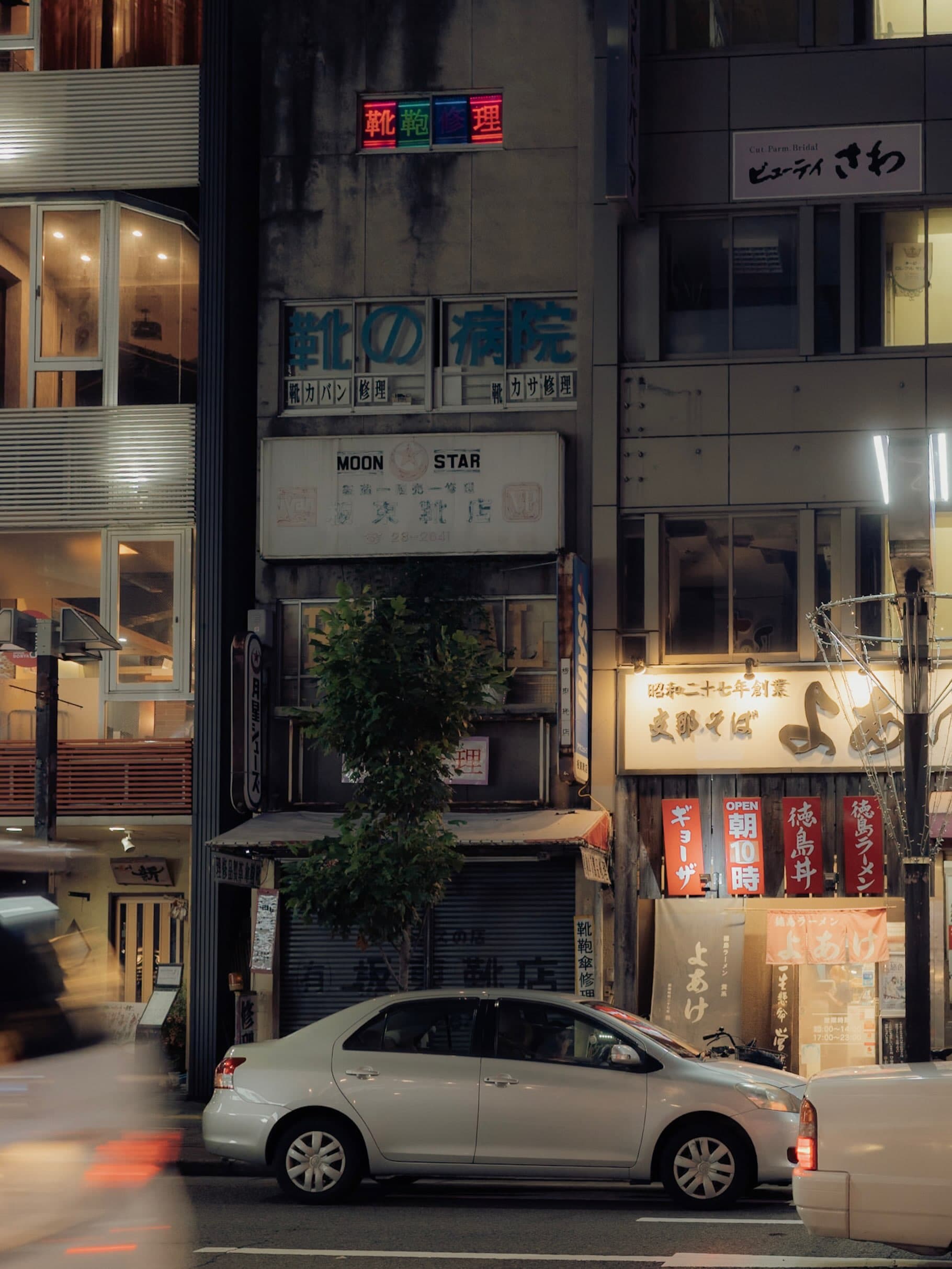夜の街角のビル。「靴の病院」「靴鞄修理」のネオン看板とMOON STARの古い看板が並び、隣は徳島ラーメン店。手前の車道に停まる白い車