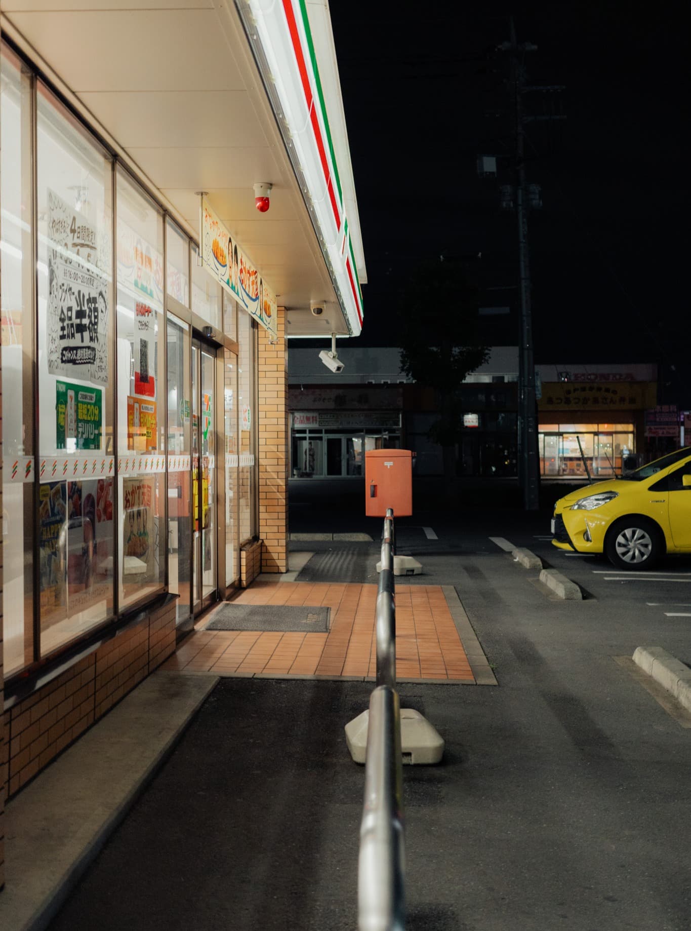 夜のコンビニ店先。窓に「全品半額」の貼り紙、歩道の先に赤いポスト、右の駐車場に黄色い車が停まる。手前に金属の手すり