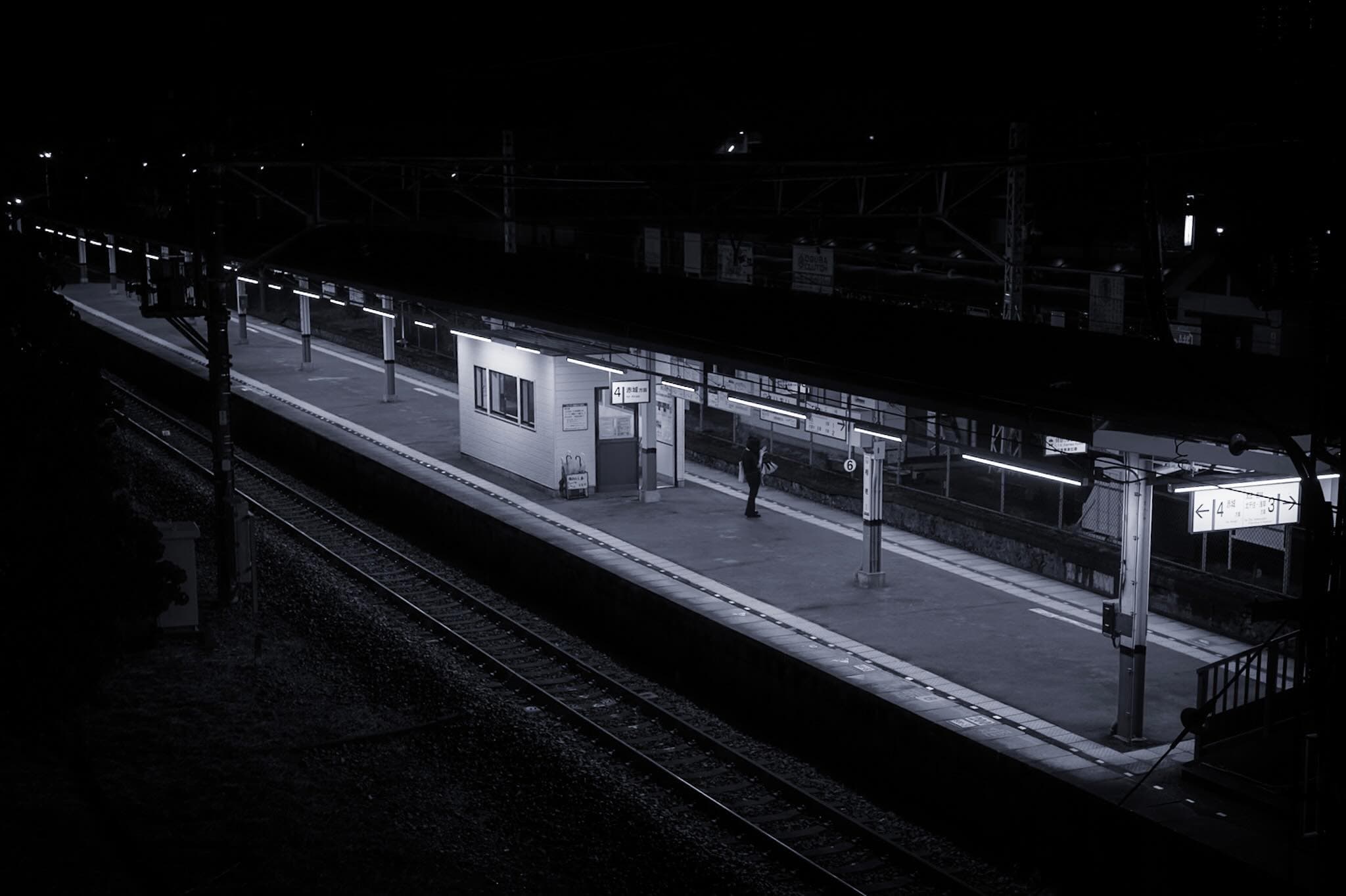 夜の駅を跨線橋から見下ろしたモノクロ。誰もいないホームに一人が立ち、駅名標に「4 赤城方面」。手前に線路と架線柱