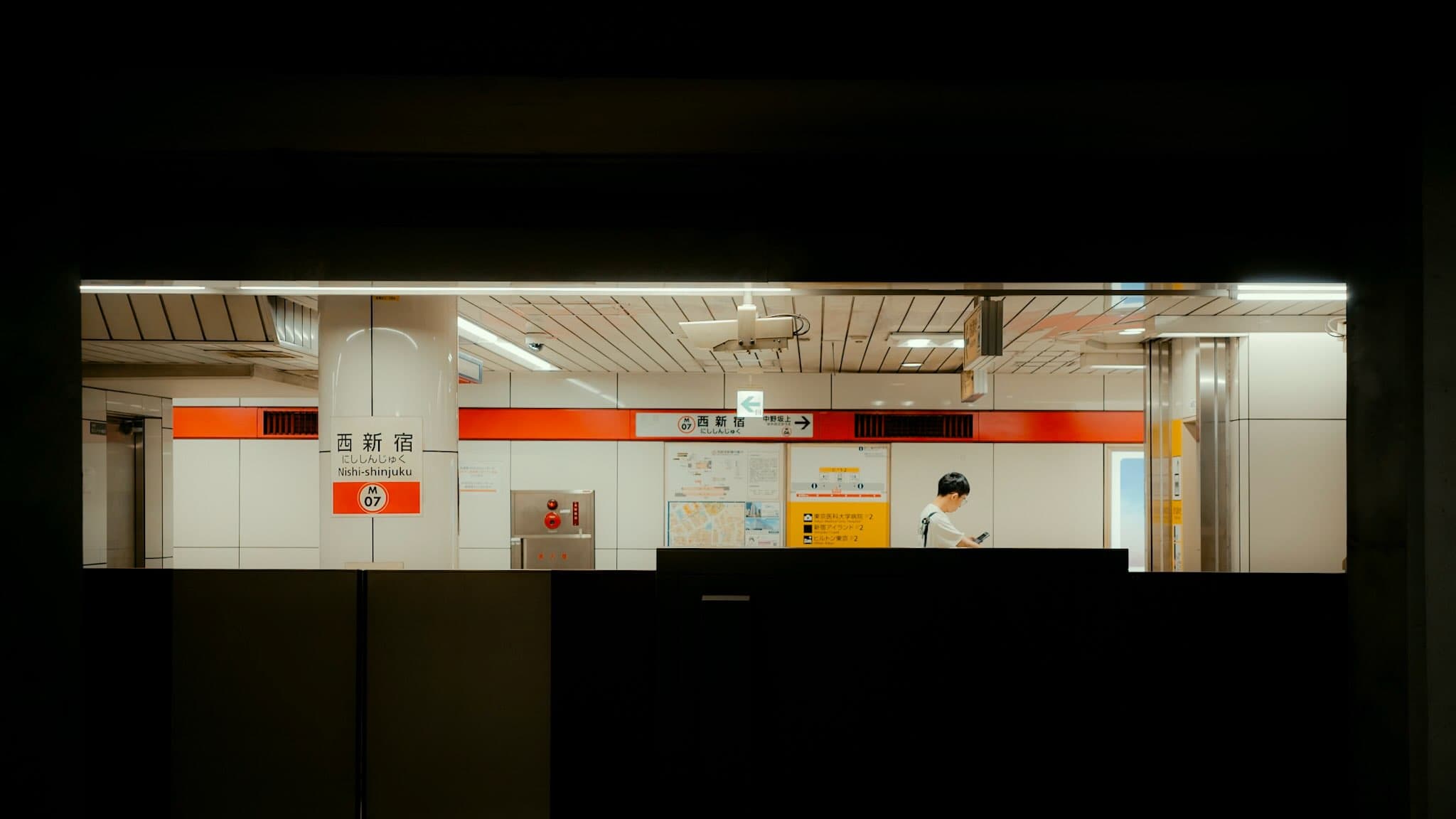 暗い枠の向こうに、西新宿（M07）の駅名標と赤い帯の壁。白いシャツの人がスマートフォンを見て立っている
