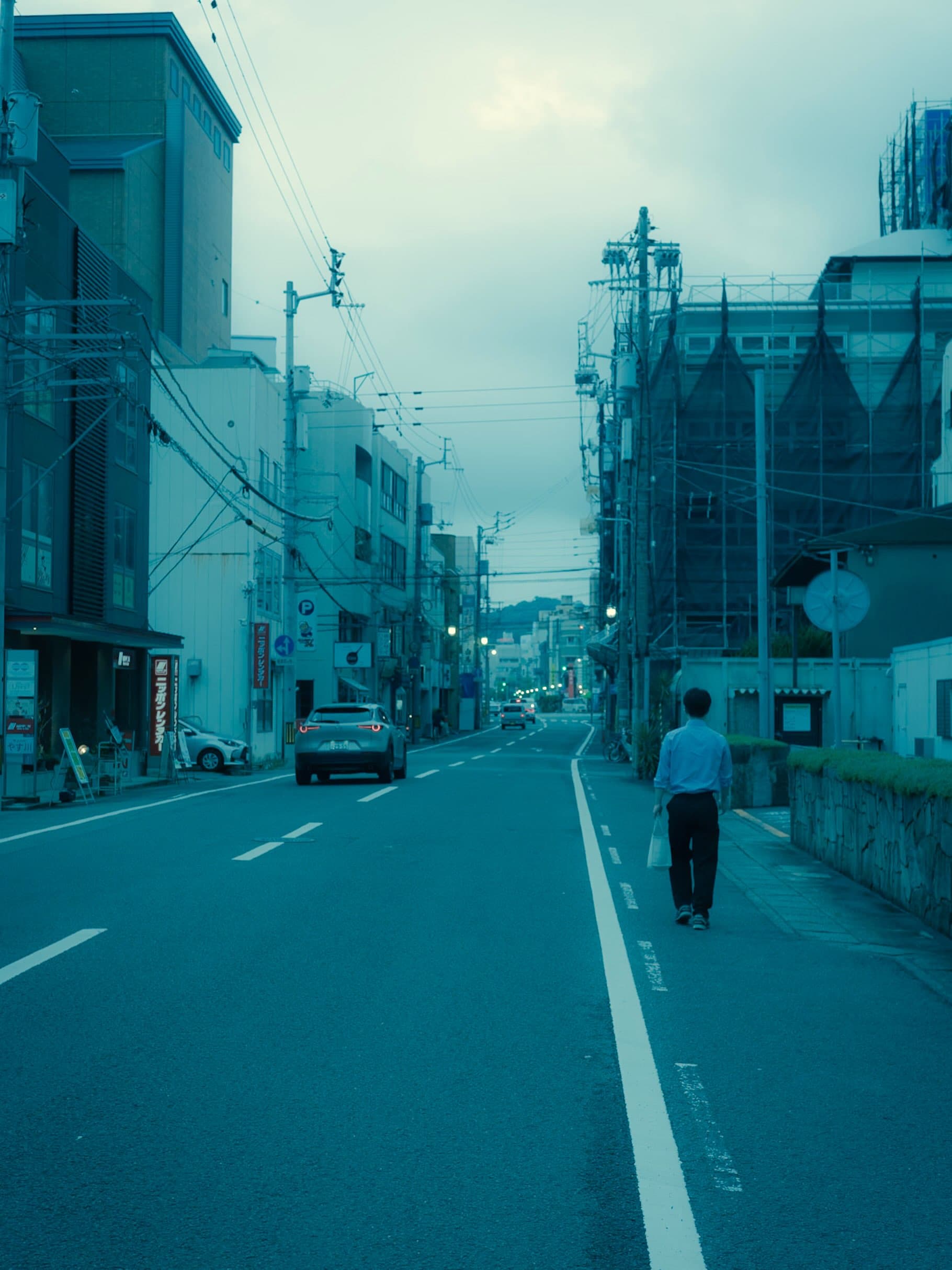 夕暮れの街路を奥へ向かって歩く白シャツの男性。車道には車が走り、電柱と電線、工事用シートを掛けた建物が並ぶ