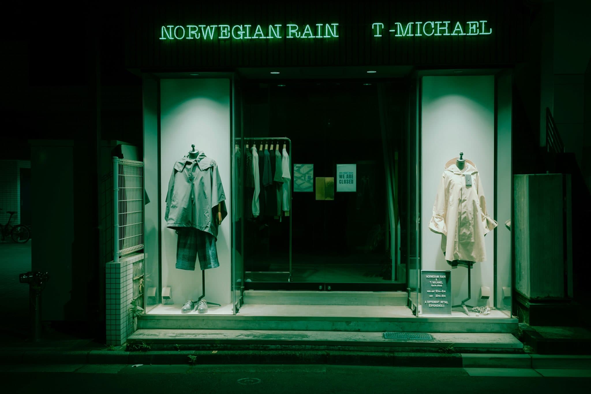 夜の店舗の外観。「NORWEGIAN RAIN」「T-MICHAEL」の緑のネオン文字が灯り、左右のショーウィンドウにコートを着せたマネキンが立つ