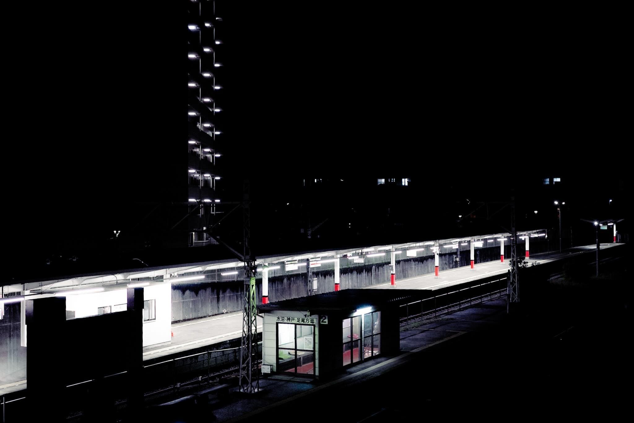 夜の無人駅ホーム。蛍光灯が一列に灯り、待合室の駅名標に「水沼・神戸・足尾方面 2」。奥に照明の並ぶ高い建物