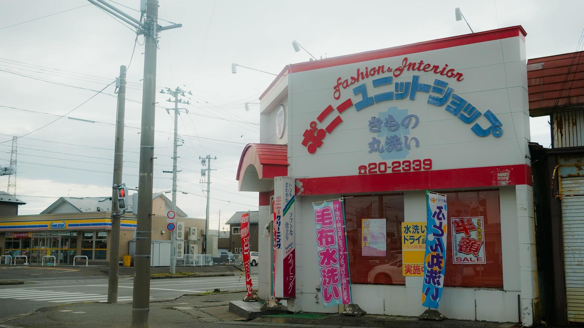 赤と白の外壁のクリーニング店。「ポニーユニットショップ」「きもの丸洗い」「20-2339」の看板と、「毛布水洗い」「半額SALE」ののぼりが並ぶ
