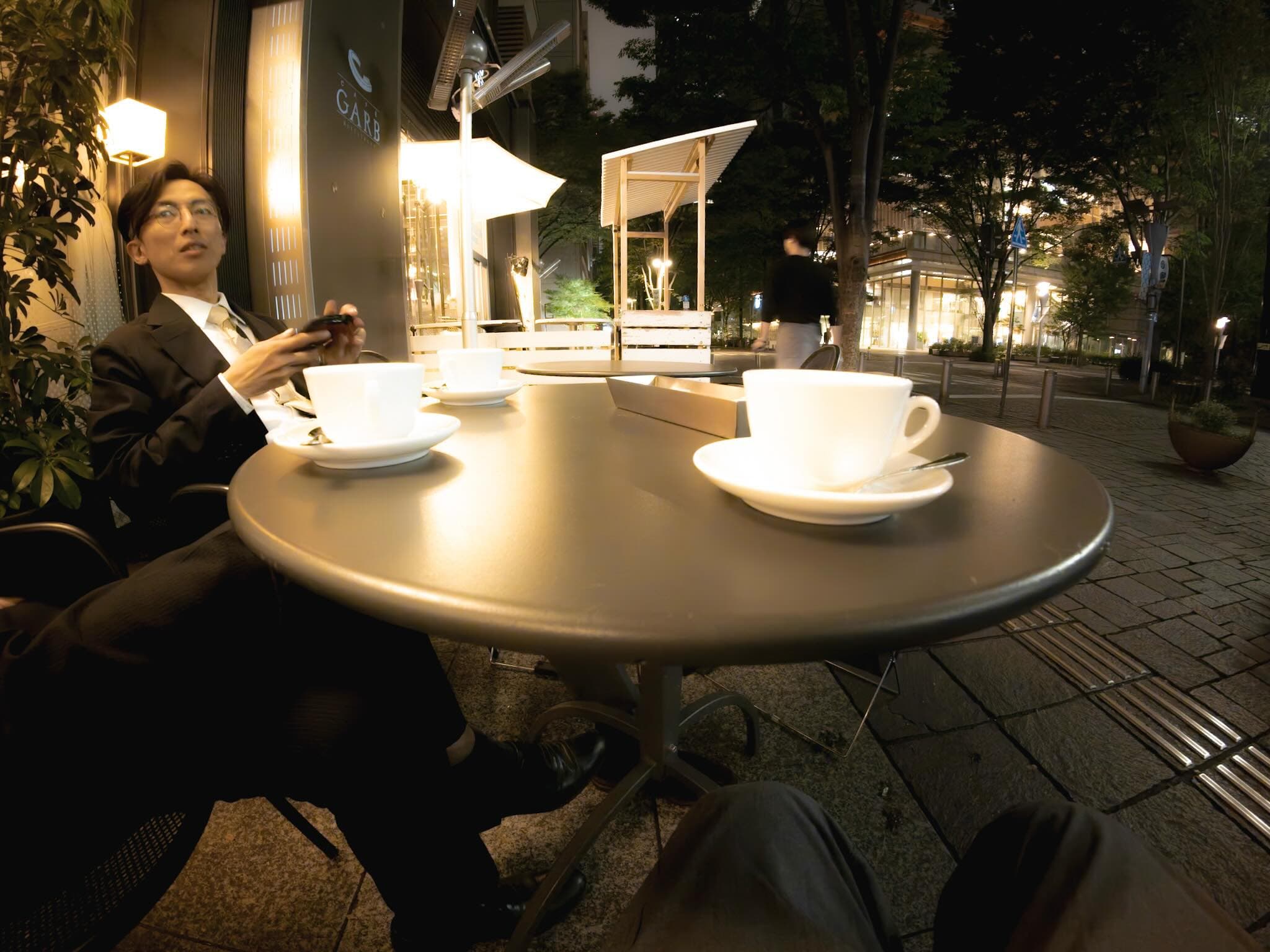 夜のカフェテラス。丸いテーブルに白いカップとソーサーが二つ置かれ、スーツ姿の男性がスマートフォンを手に座る。奥の柱に「GARB」の看板