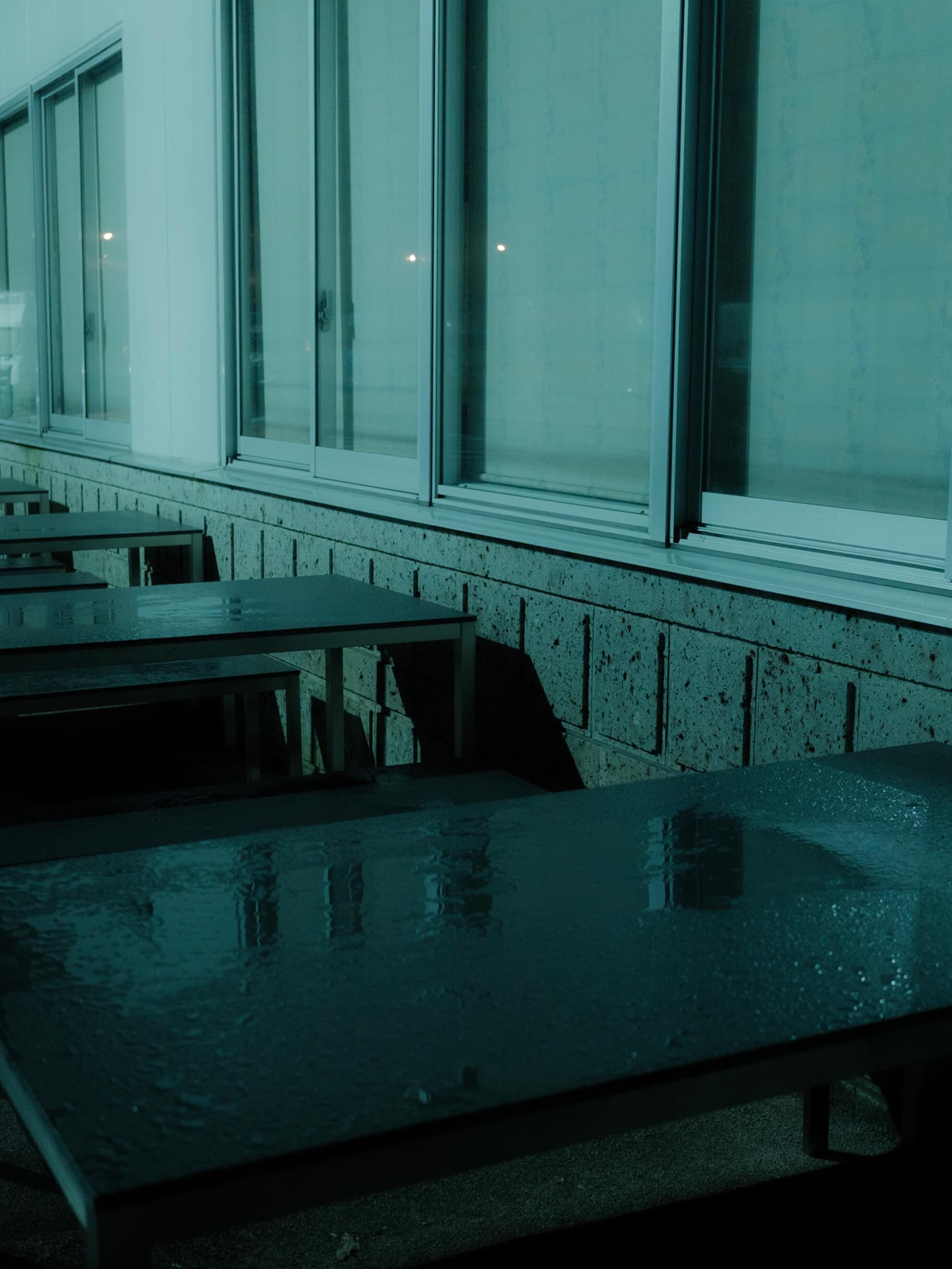 夜のガラス張りの建物の外、雨に濡れて水を張ったようになった屋外テーブルが並ぶ。壁は穴の空いた石張り、全体が青緑に沈む