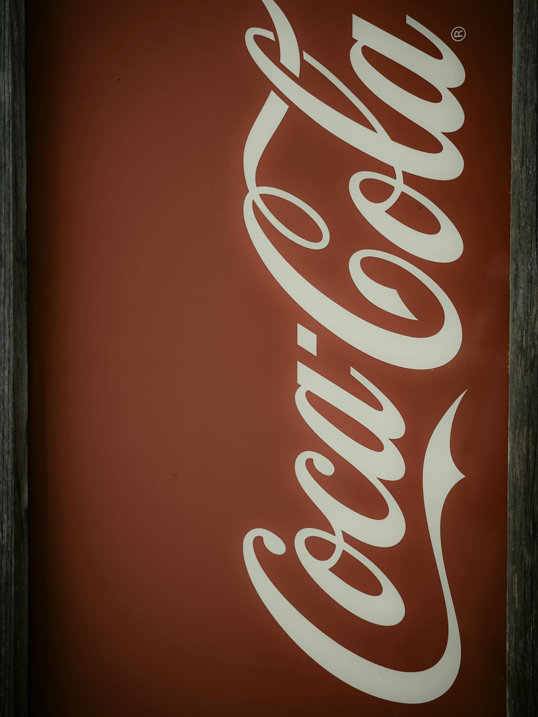 古びた木枠にはめ込まれた赤地のCoca-Cola看板。白い筆記体ロゴが縦に大きく入り、右上に登録商標マーク