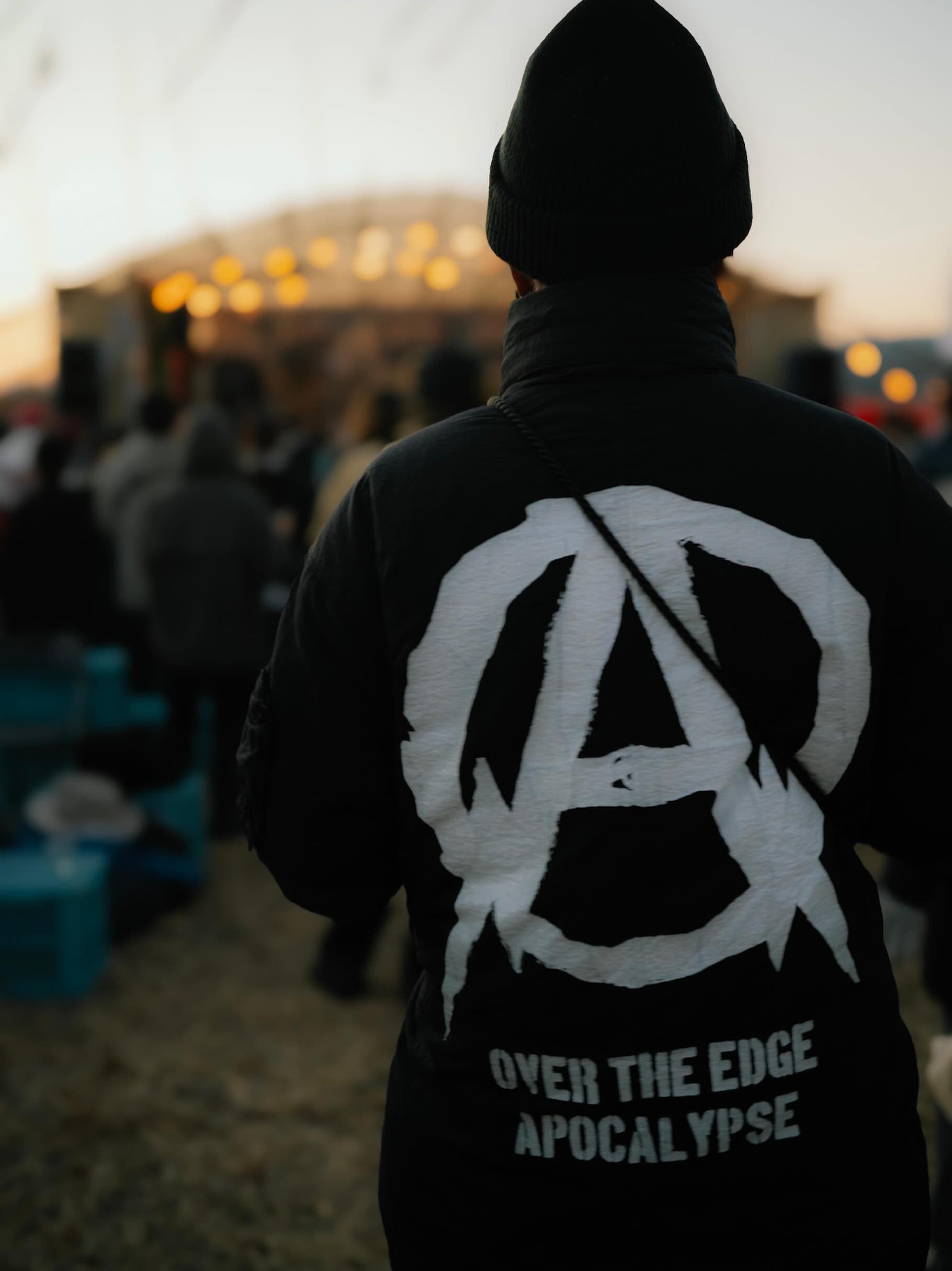 ニット帽と黒いジャケットの後ろ姿。背中にアナキストマークと「OVER THE EDGE APOCALYPSE」の白い文字。奥に照明の灯る野外ステージと観客