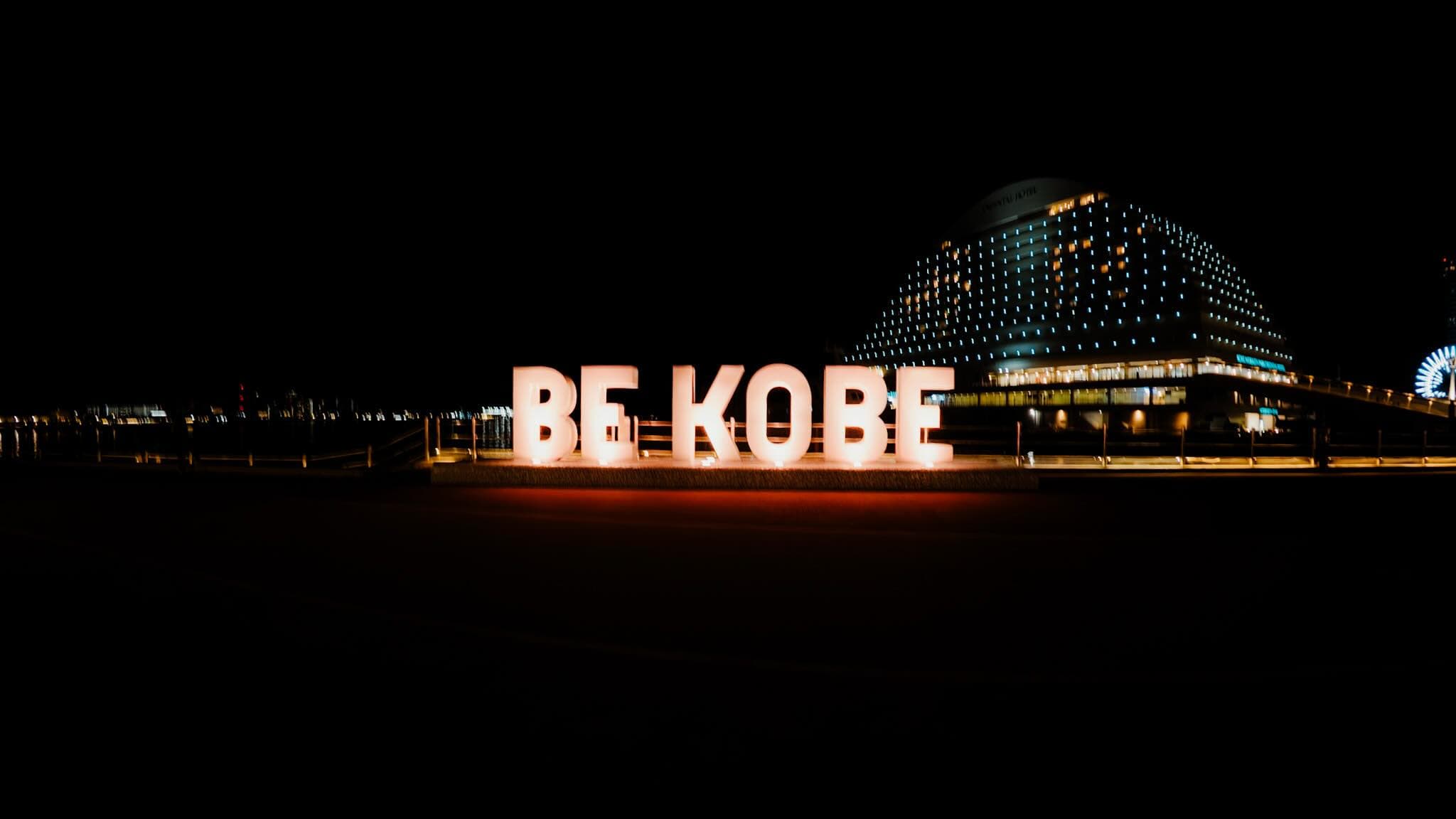 夜の海辺に「BE KOBE」の白い立体文字が光り、奥にホテルの建物と観覧車の明かりが見える