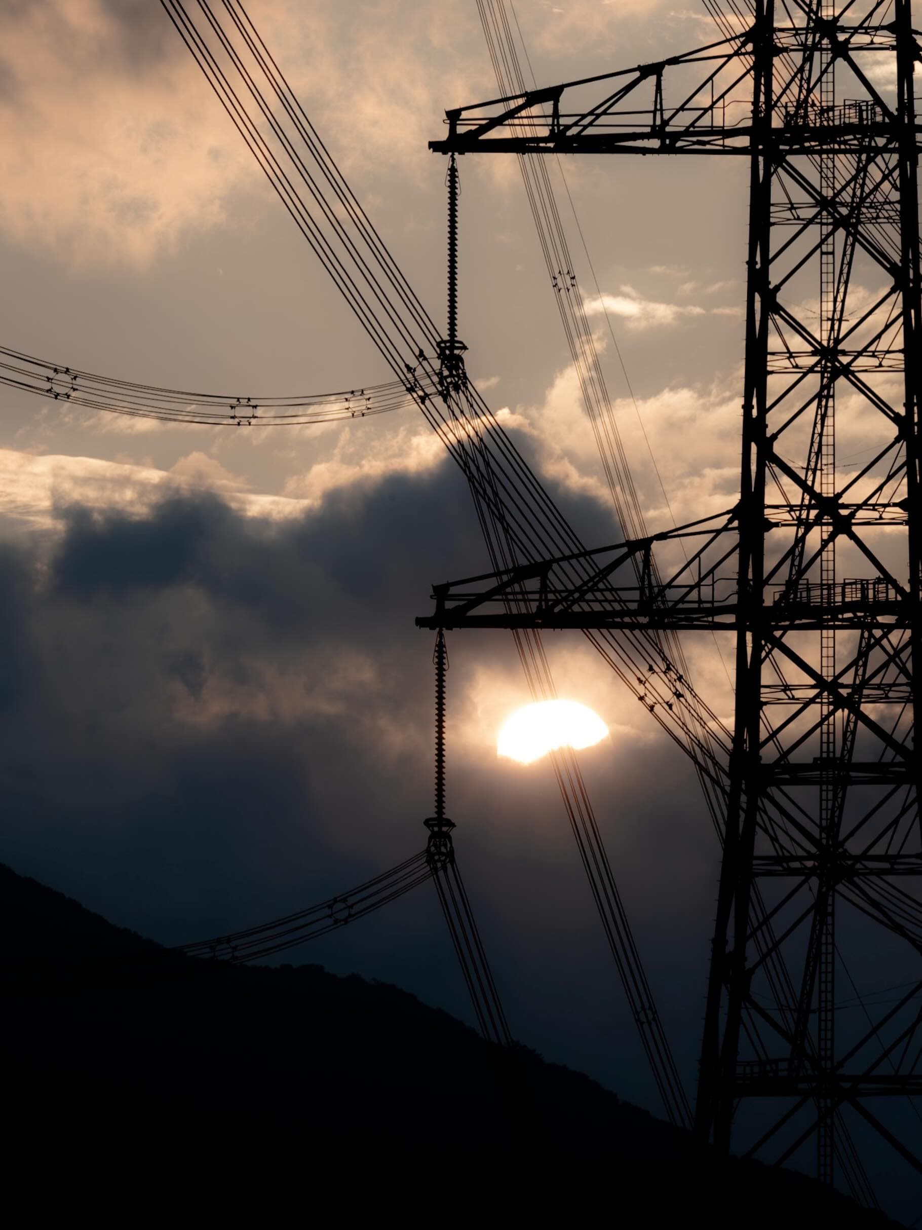 夕日を背にした送電鉄塔のシルエット。雲間から太陽がのぞき、碍子と何本もの電線が斜めに横切る。左下に山の稜線