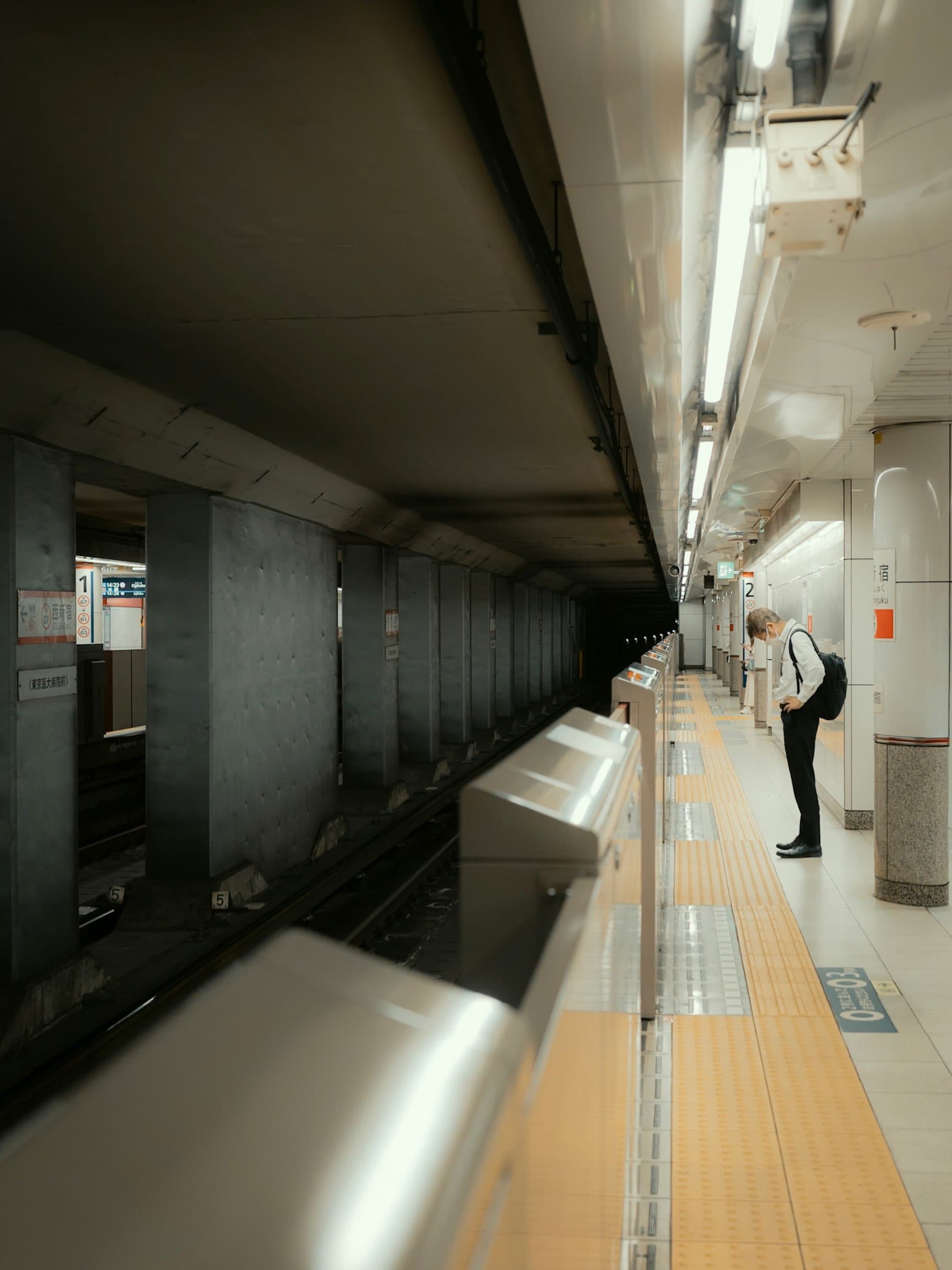 地下鉄西新宿駅のホーム。左に線路とホームドアが続き、右の黄色い点字ブロックの脇でリュックを背負った人が下を向いて立つ