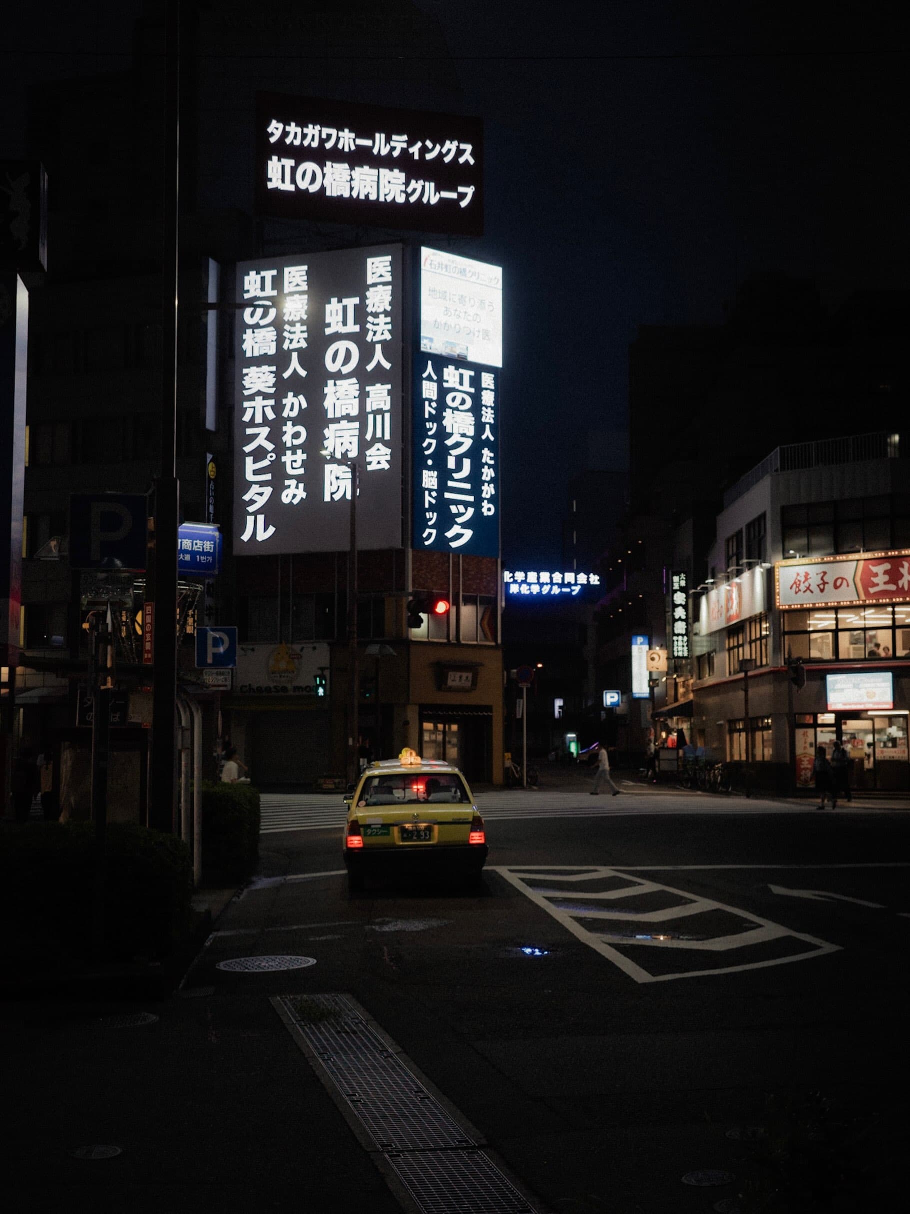 夜の交差点に停まる黄色いタクシー。頭上に「虹の橋病院」「虹の橋クリニック」の白い電飾看板、右奥に餃子の王将の店舗が光る