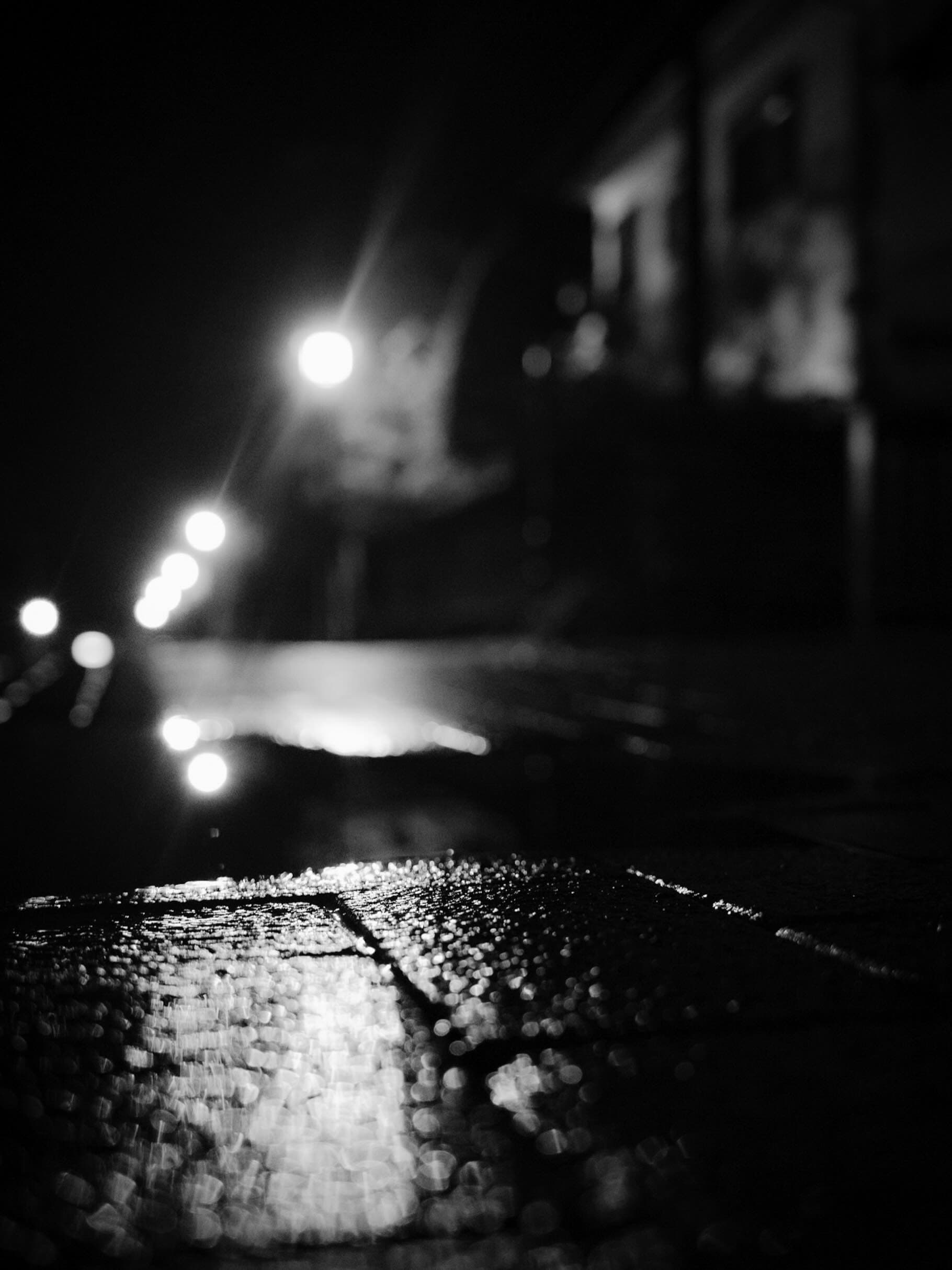 雨の夜、路面すれすれの低い位置から見た街路。奥へ続く街灯の玉ボケが並び、手前の濡れた敷石が光を反射するモノクロ