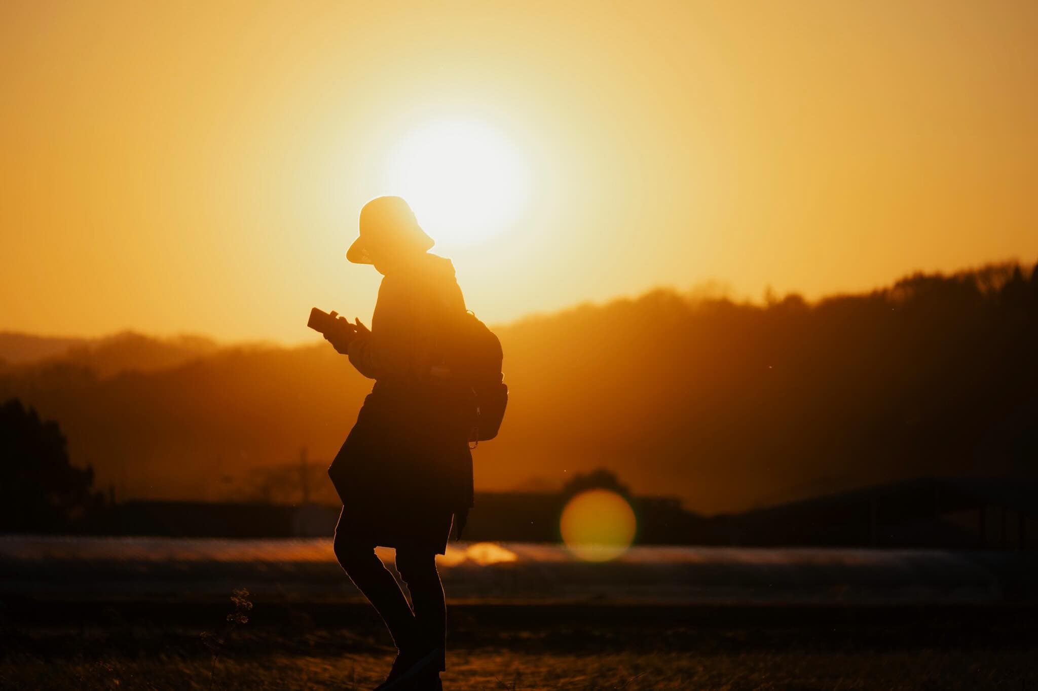 オレンジ色の夕日を背にしたシルエットの人物。ハットをかぶりリュックを背負い、両手でカメラを構えている。奥に低い山並み