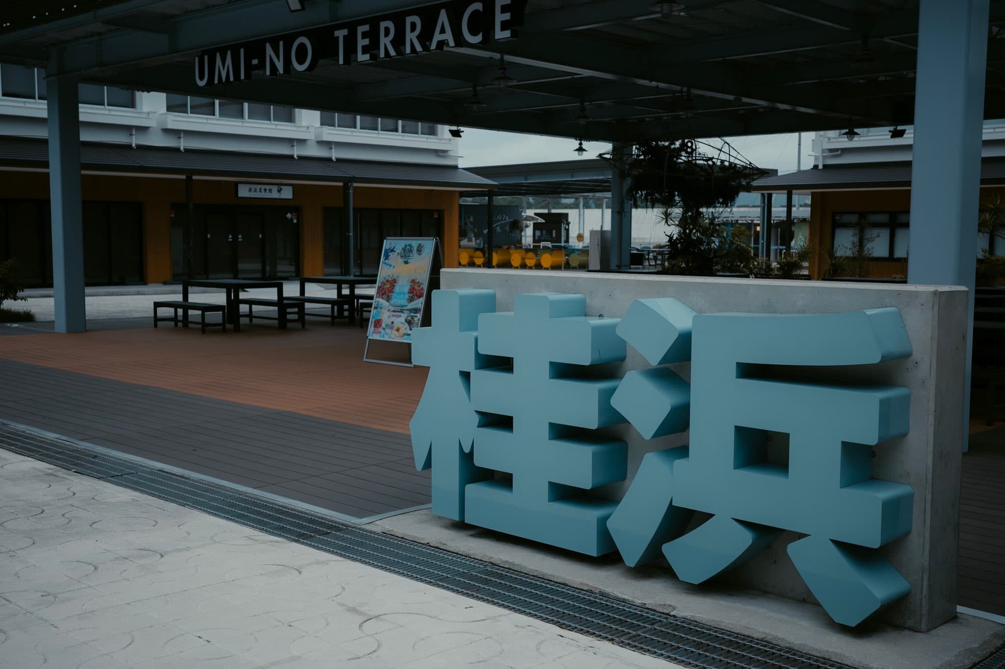 「UMI-NO TERRACE」の看板が掛かる屋根の下、コンクリート壁に水色の立体文字「桂浜」が据えられている。人のいないデッキとベンチ