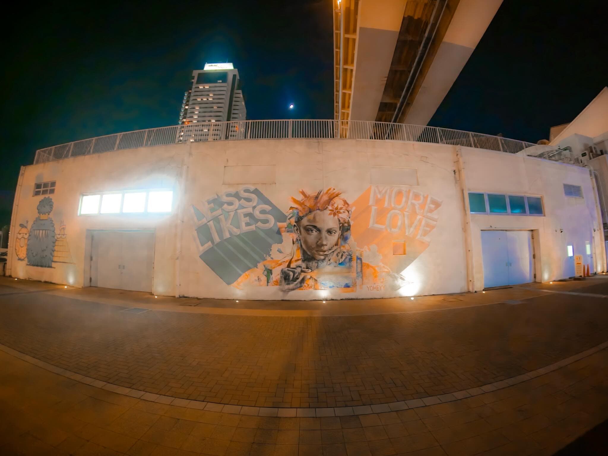 高架下の白い壁に描かれた壁画。「LESS LIKES MORE LOVE」の文字と人物の顔が並び、足元のライトに照らされている。奥に高層マンションと月