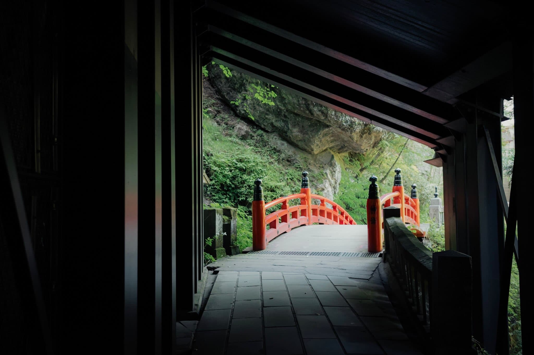 暗い建物の中から見た、朱塗りの欄干の反り橋。奥は苔むした岩壁と濃い緑の草木で、手前は石畳の通路