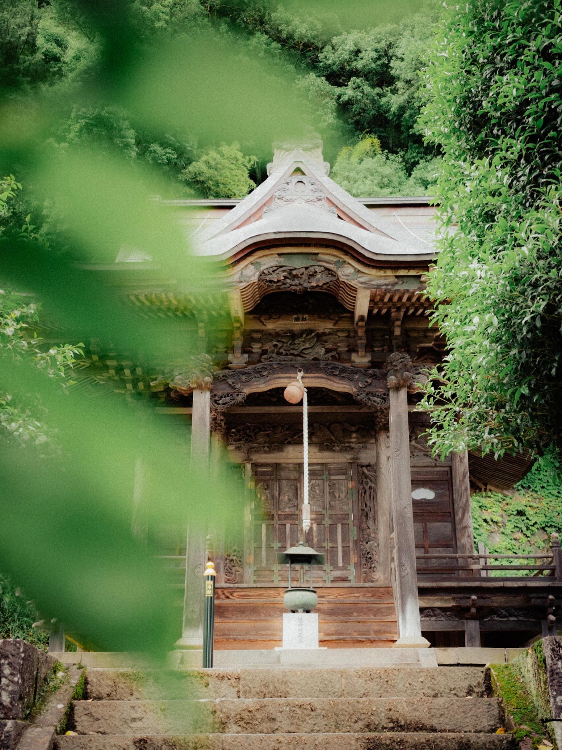山の斜面の緑に囲まれた木造のお堂。彫刻の入った向拝から鈴緒が下がり、手前の石段に香炉が据えられている