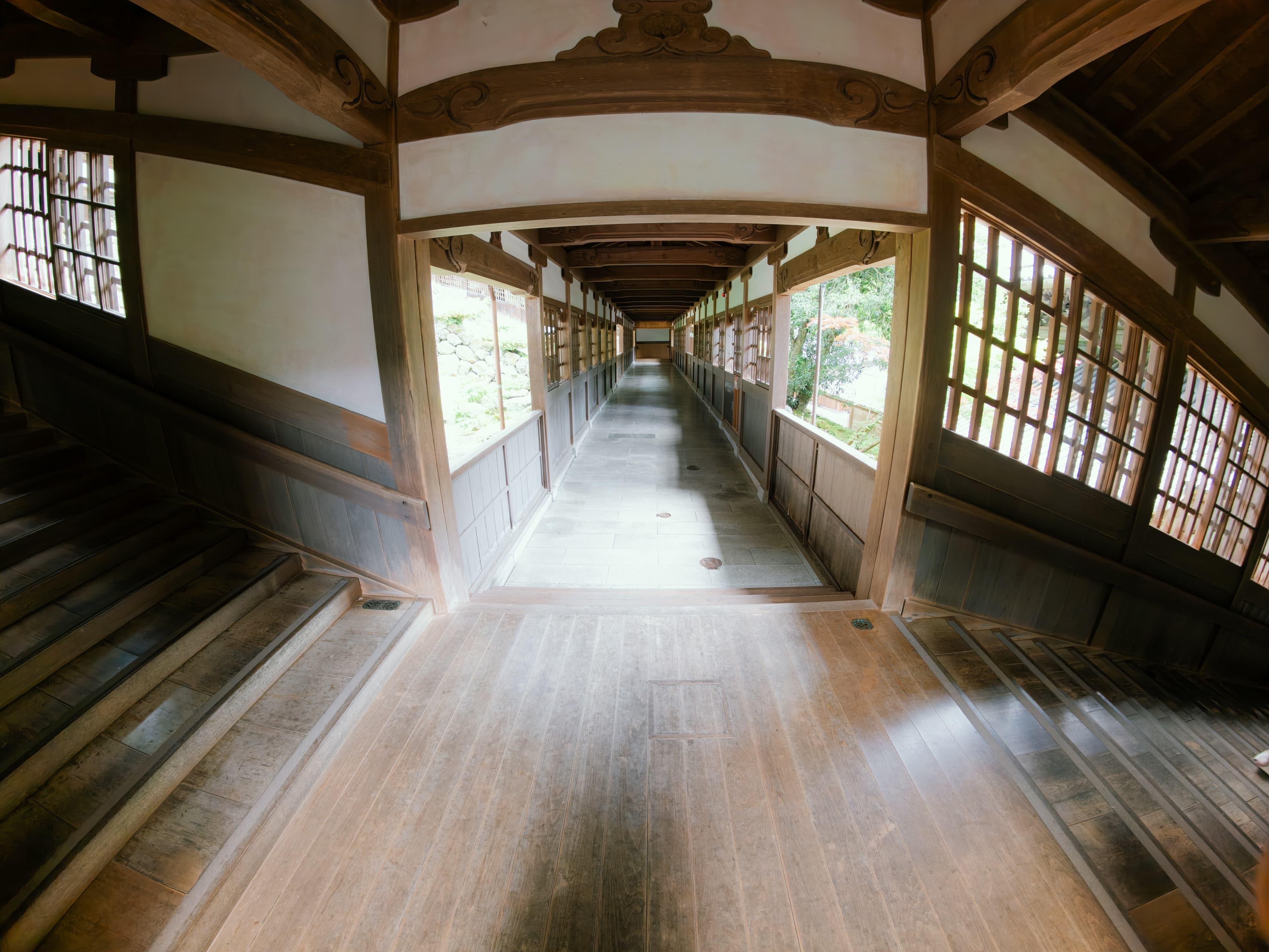 木造の回廊を見下ろした眺め。中央の板張りの通路が奥へまっすぐ延び、左右に階段と格子窓、窓の外に石垣と庭木が見える