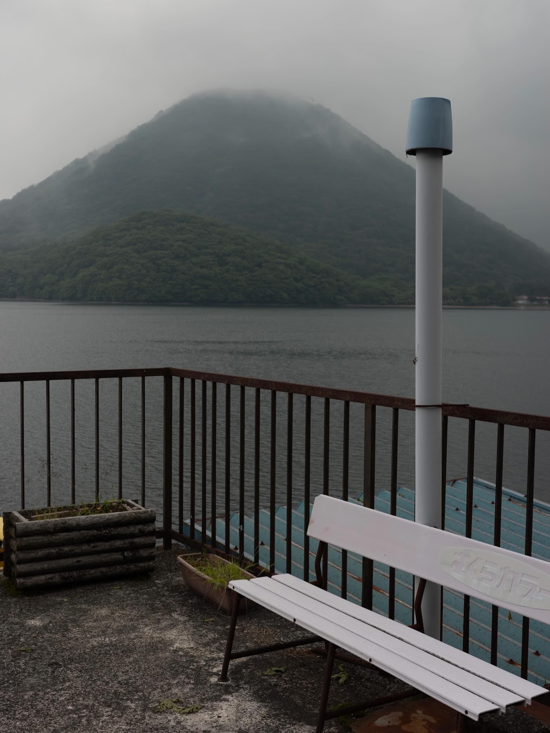 曇り空の湖畔、錆びた柵の内側に白い木製ベンチ。対岸には霧のかかった円錐形の山、手前に草の生えた古いプランター