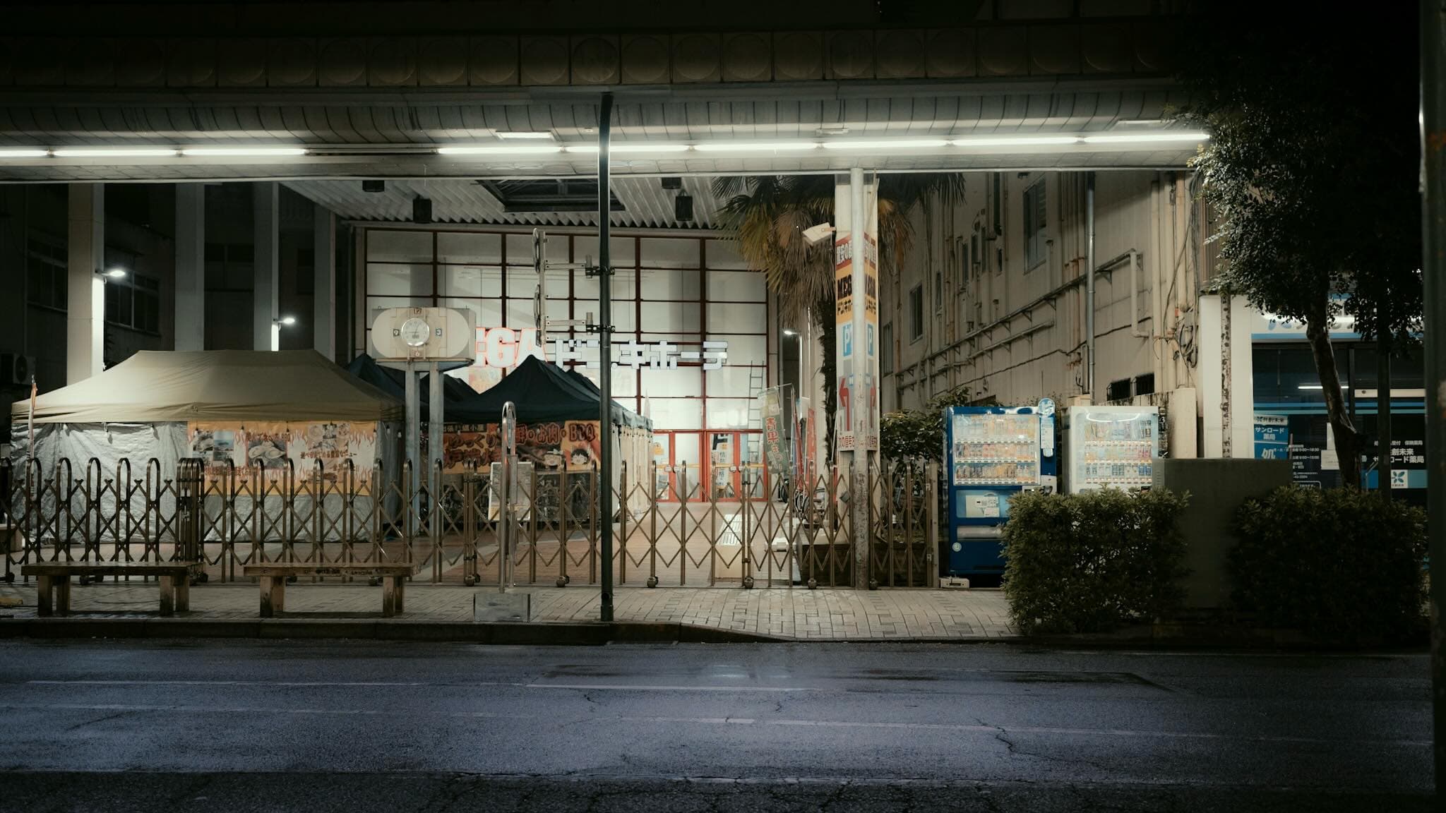 夜のMEGAドン・キホーテ店頭。閉店後の蛇腹門が閉まり、テント屋台と自動販売機が並ぶ前を蛍光灯が照らしている