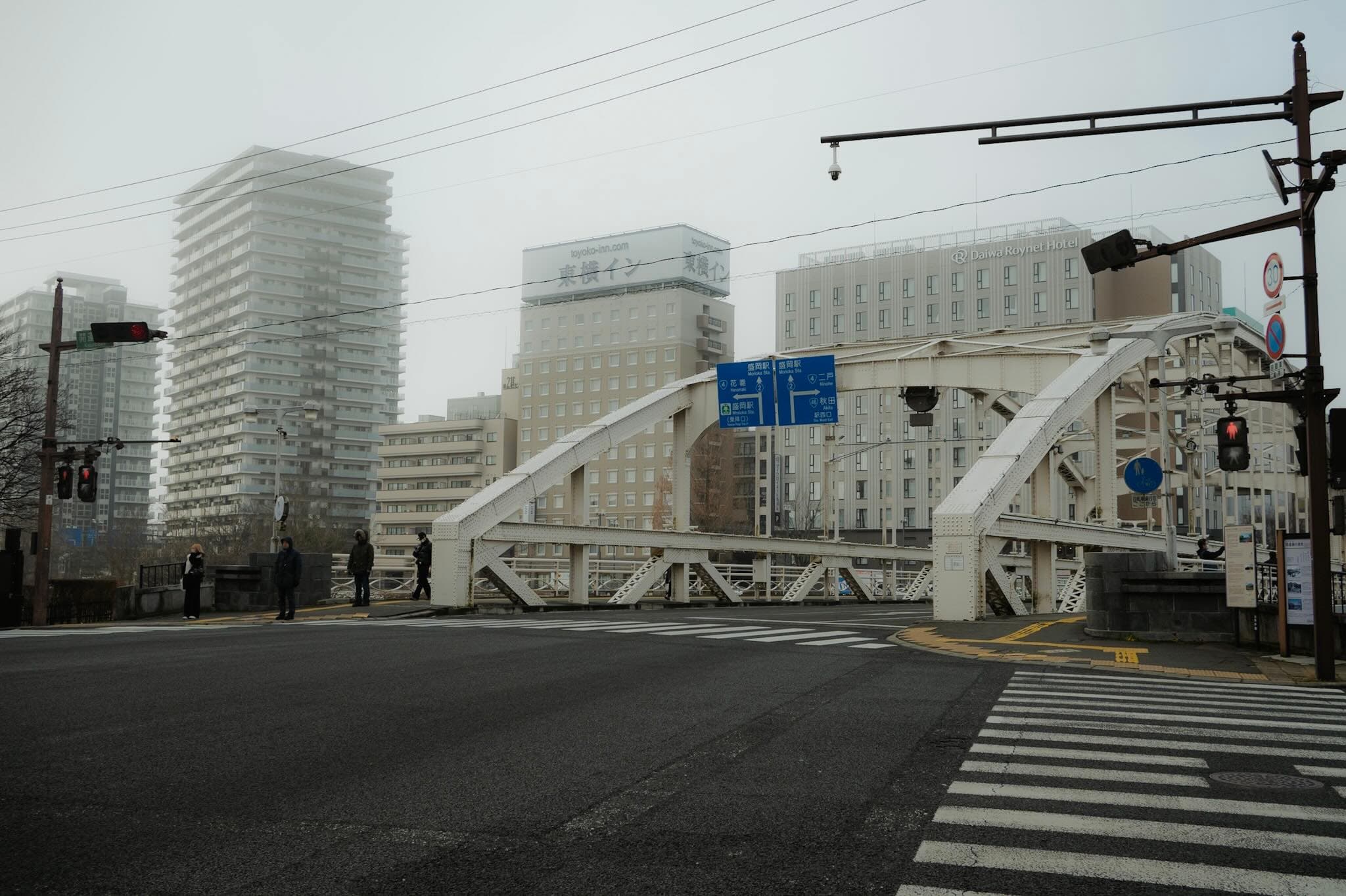 曇り空の交差点。白い鉄骨アーチ橋の手前に横断歩道と信号があり、青い案内標識に「盛岡駅」「花巻」「二戸」の表示。奥に高層マンションとホテル