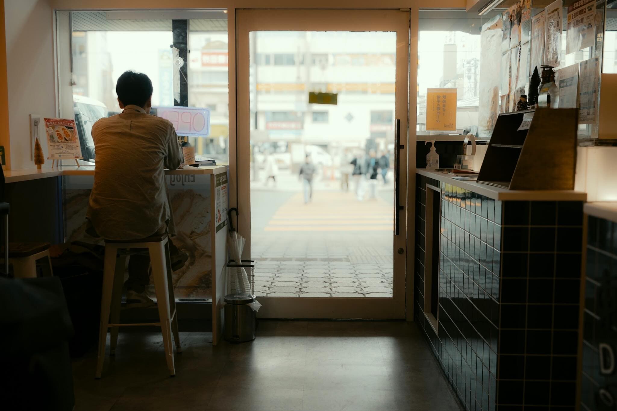 店内から見た通りへのガラス戸。手前でスツールに座った男性が窓際のカウンターに向かい、外の横断歩道を人が歩いている
