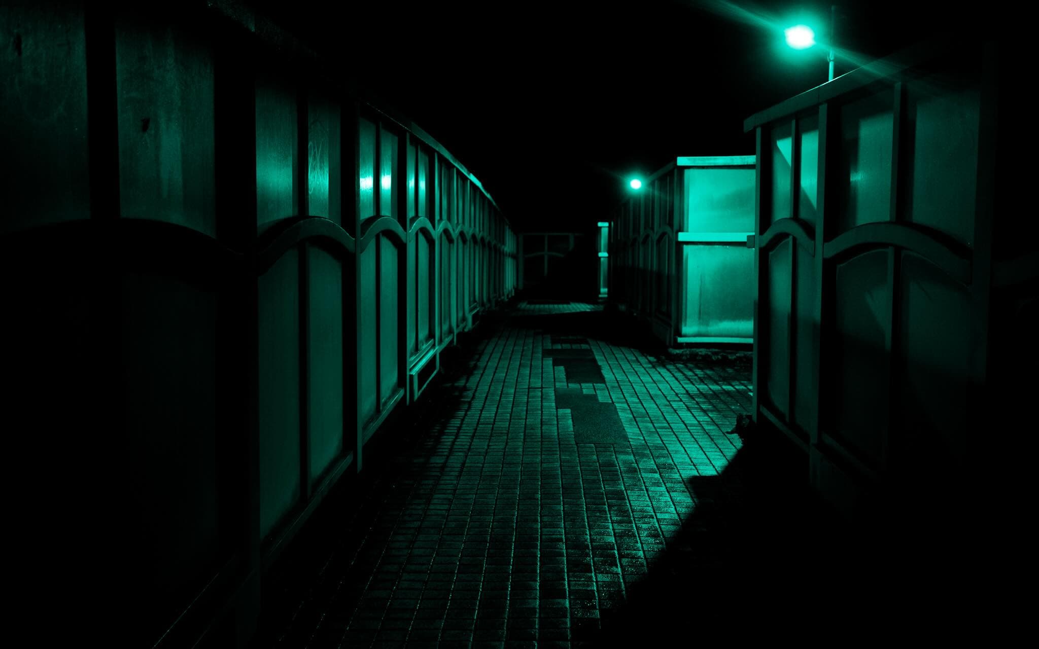 夜の煉瓦敷きの通路。両側に曲線の意匠が入った塀が続き、街灯二基が通路を青緑に照らす