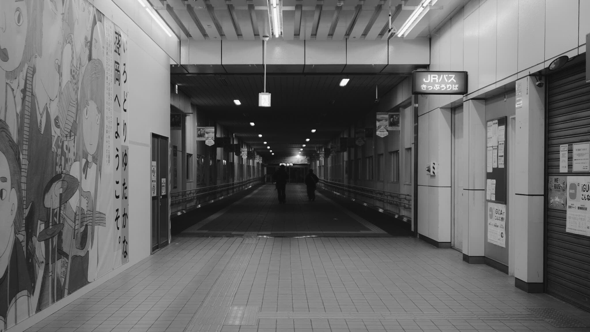 モノクロの駅地下通路。左壁に「いろどりゆたかな盛岡へようこそ。」の垂れ幕とイラスト、右奥に「JRバス きっぷうりば」の看板。奥を2人が歩く
