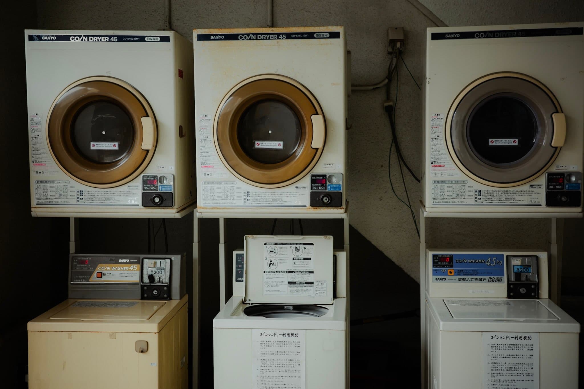 コインランドリーに並ぶSANYOの古い乾燥機三台と、その下に置かれた洗濯機。壁には「コインランドリー利用規約」の紙が貼られている
