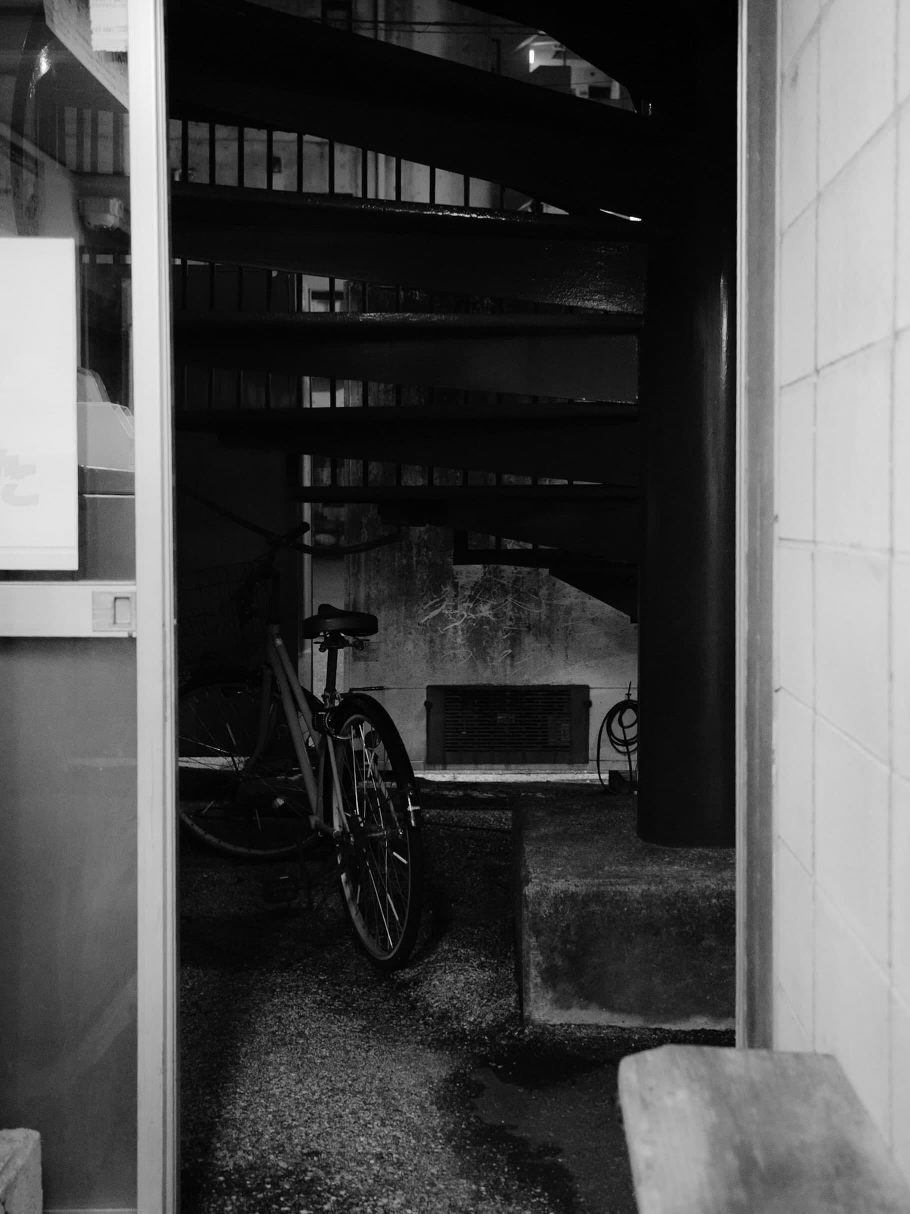 建物の隙間の暗い通路に自転車が一台停められ、奥に階段の裏側とコンクリートの壁が見えるモノクロの光景