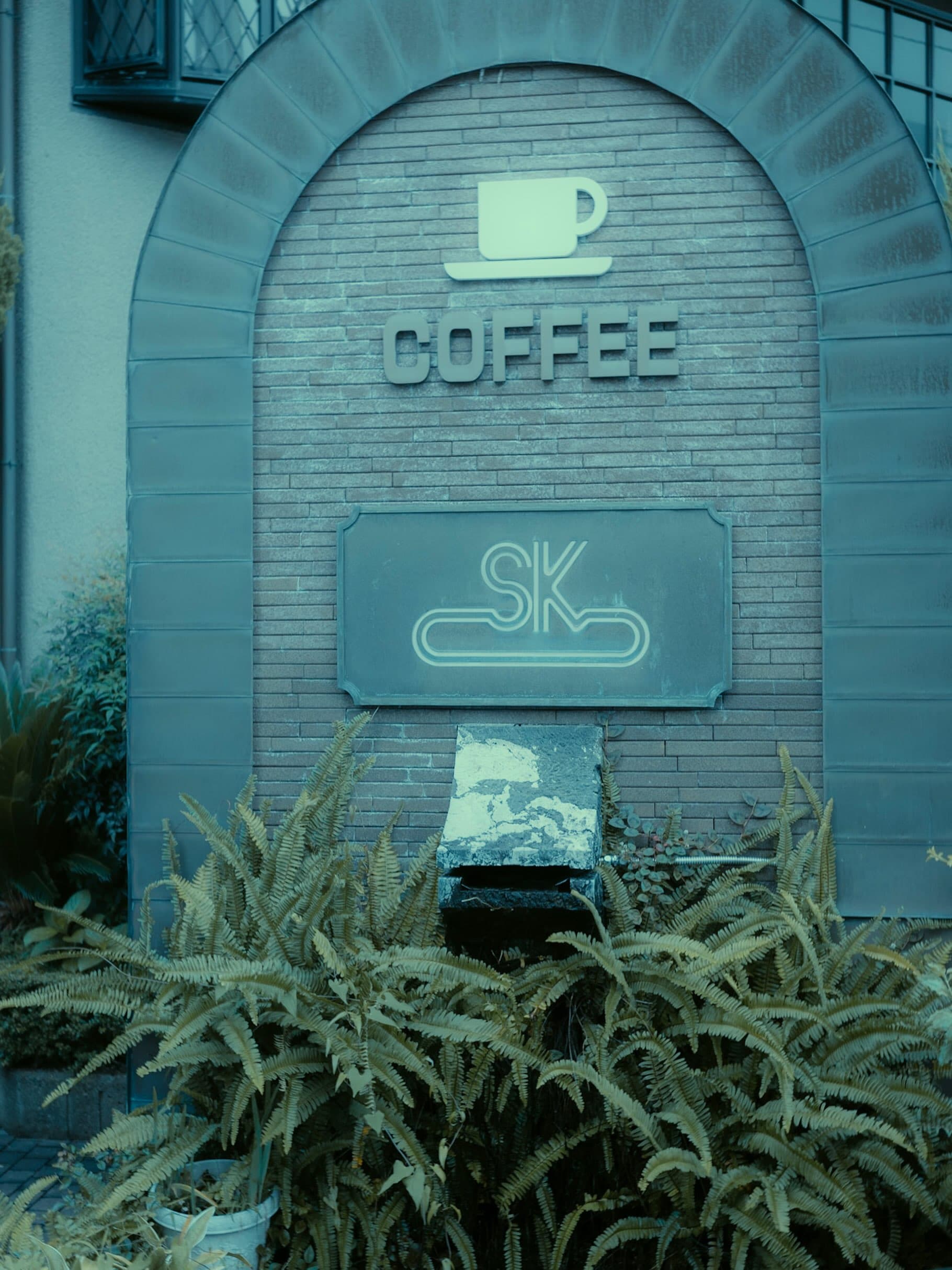 アーチ形のレンガ壁に、カップのマークと立体文字の「COFFEE」、SKのロゴ板が取り付けられている。手前にシダが茂り、全体が青緑がかった色