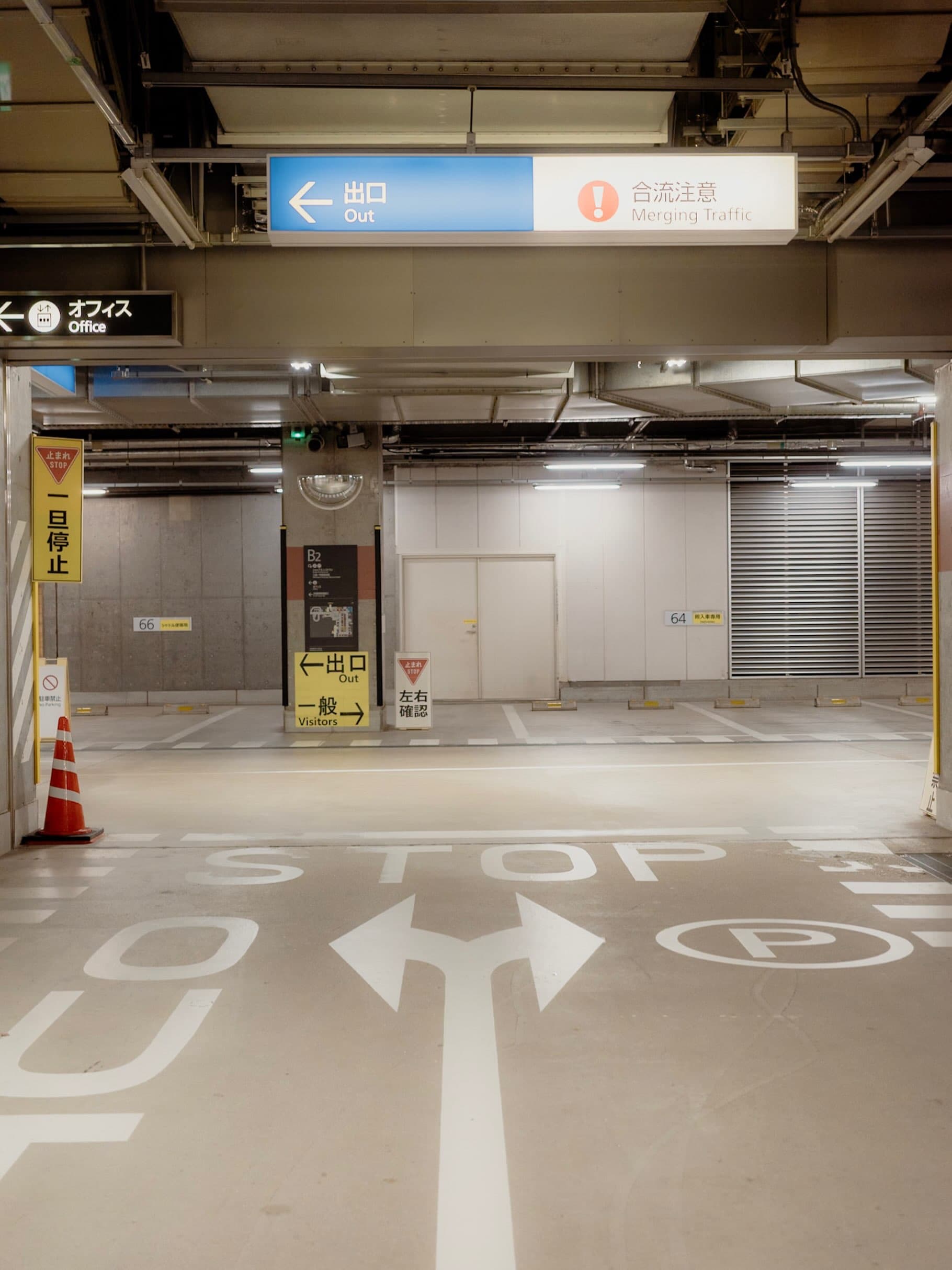 地下駐車場。「出口 Out」「合流注意 Merging Traffic」の案内板が下がり、床には白いSTOPの文字と分岐する矢印、脇に赤いコーンが1つ