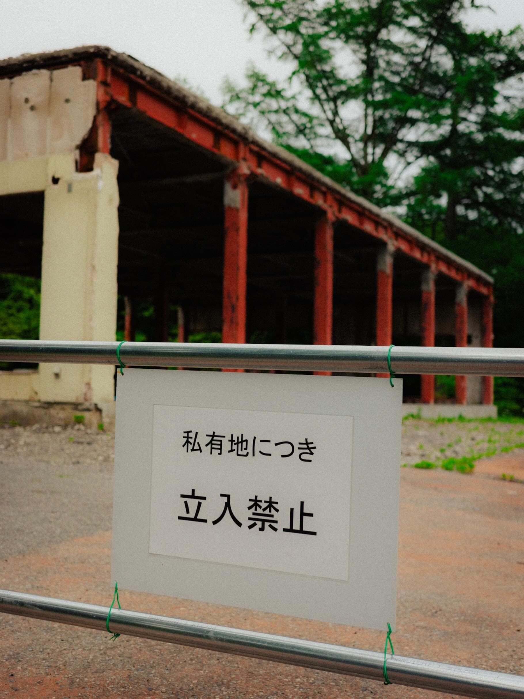 単管パイプの柵に「私有地につき立入禁止」の白い札が結ばれ、奥に赤い鉄骨の柱が並ぶ廃屋が見える