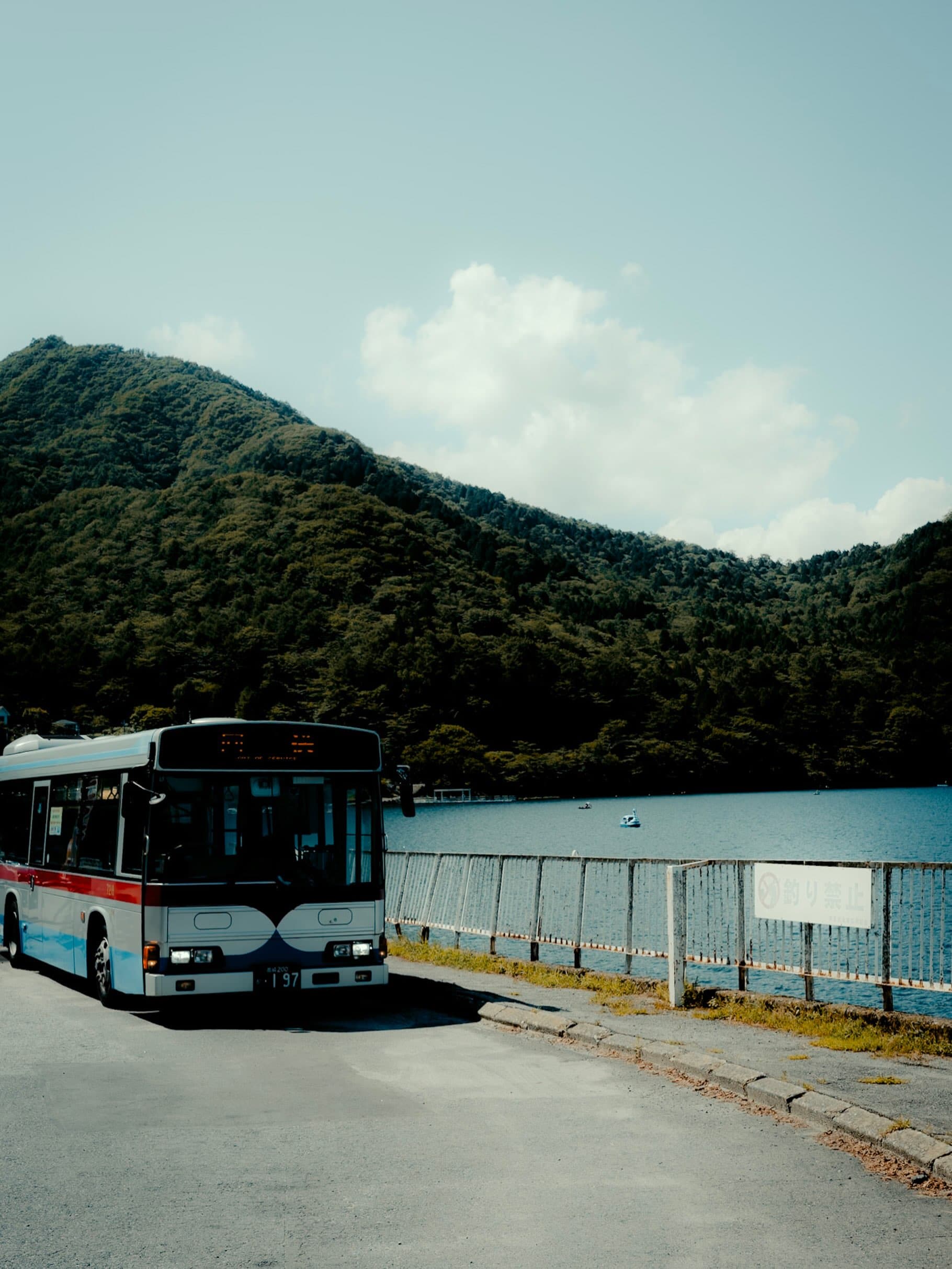 湖畔の道路に停まる白と赤の路線バス。背後に緑の山が連なり、柵の向こうの湖に白鳥型ボートが浮かび、「釣り禁止」の看板が立つ