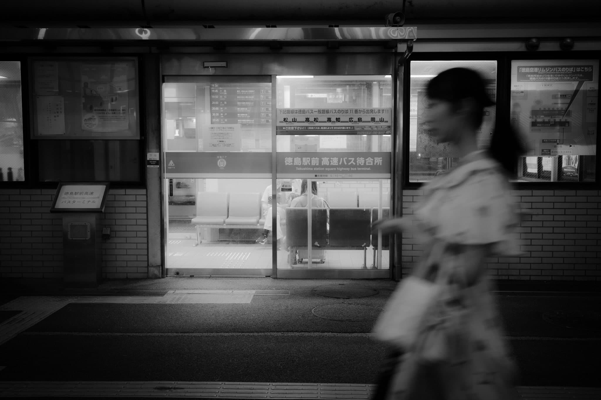 「徳島駅前 高速バス待合所」のガラス扉。中のベンチに座る人が見え、手前を手荷物を持った人物が横切ってぶれて写るモノクロ