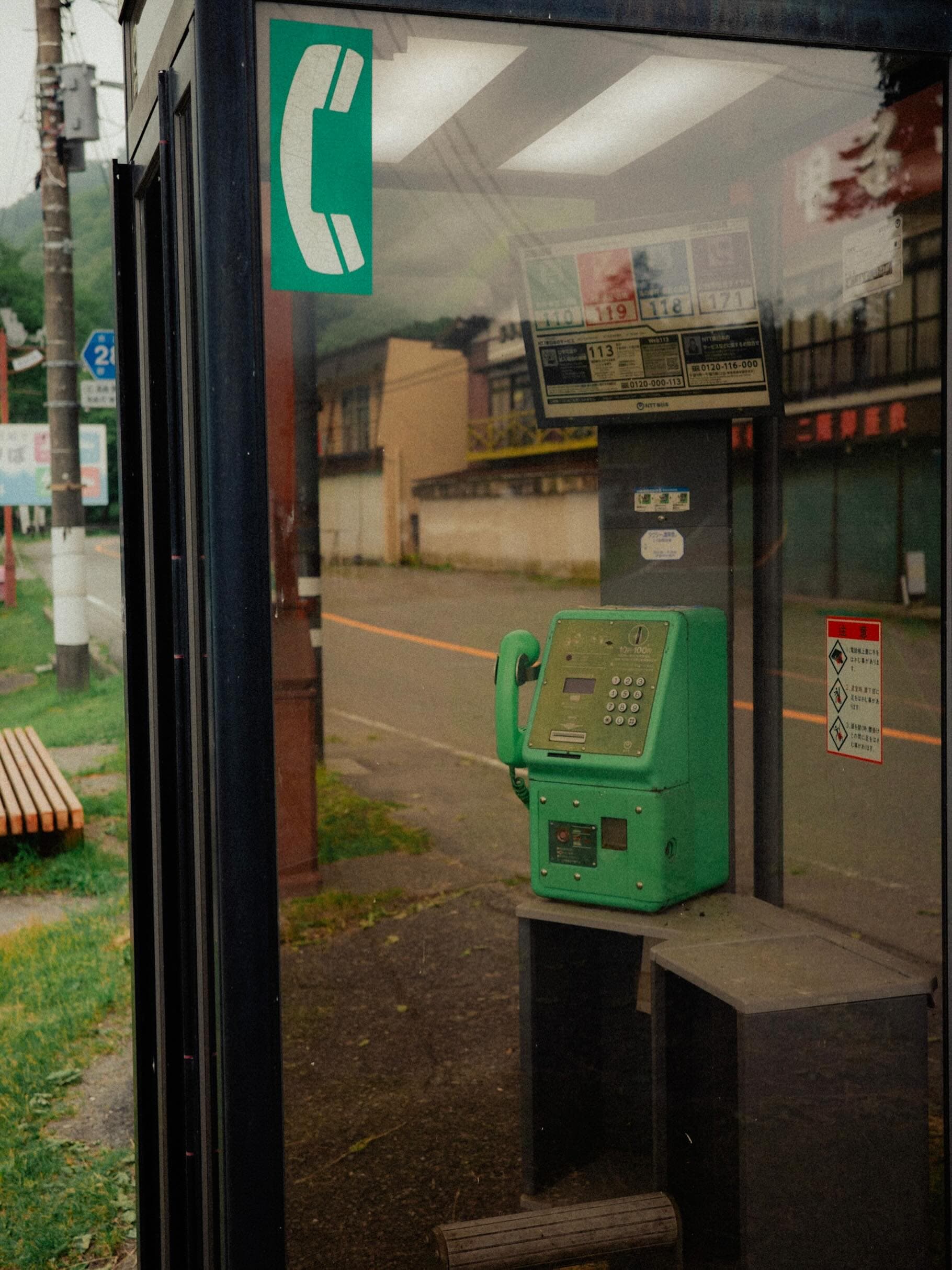 公衆電話ボックスのガラス越しに緑の公衆電話が写り、110・119・171の案内板が掲げられている。ガラスには道路と町並みが映り込む