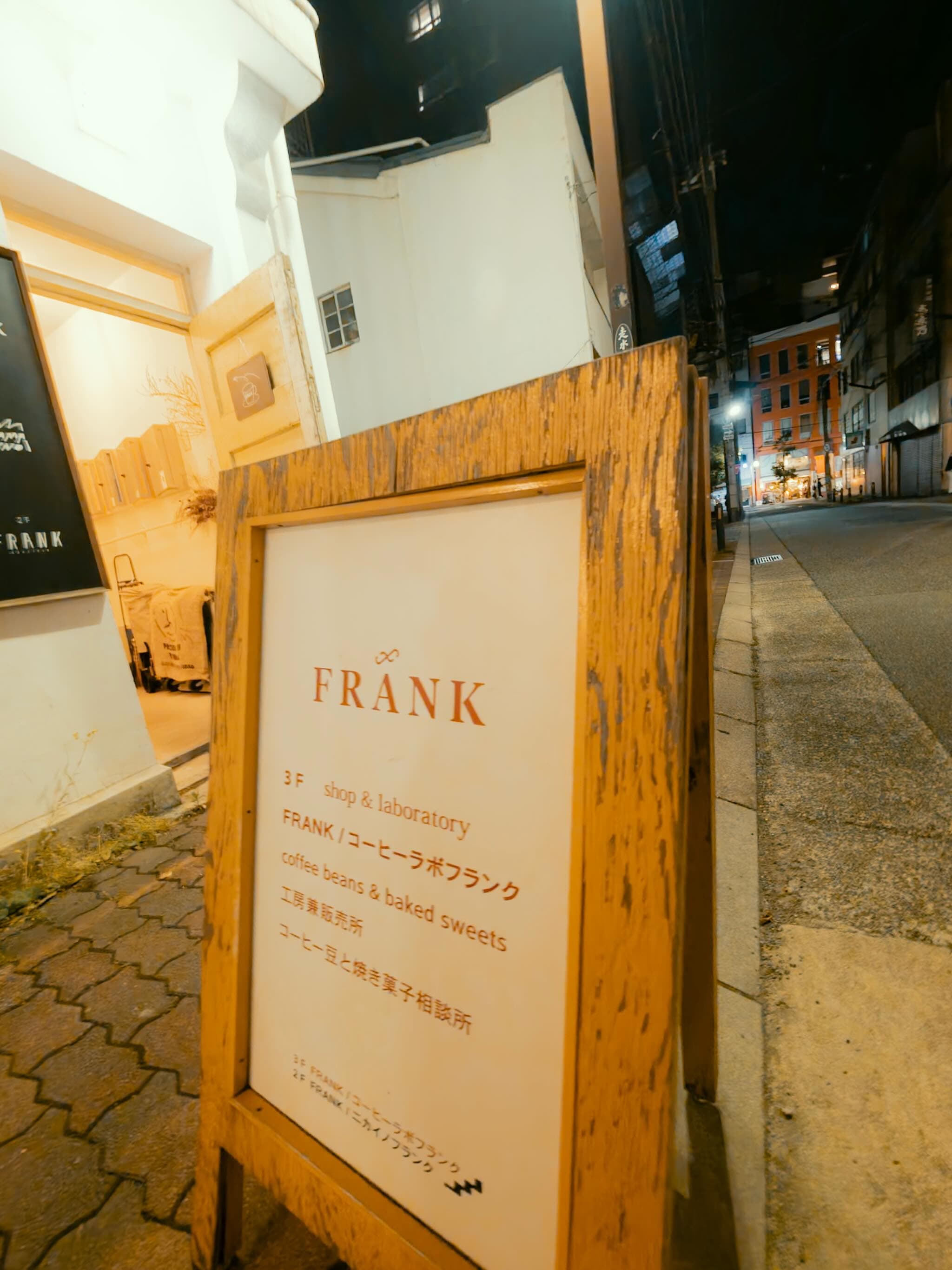 夜の路地に置かれた木枠の立て看板。「FRANK 3F shop & laboratory コーヒー豆と焼き菓子相談所」と書かれ、奥に白い建物と下り坂の道が続く