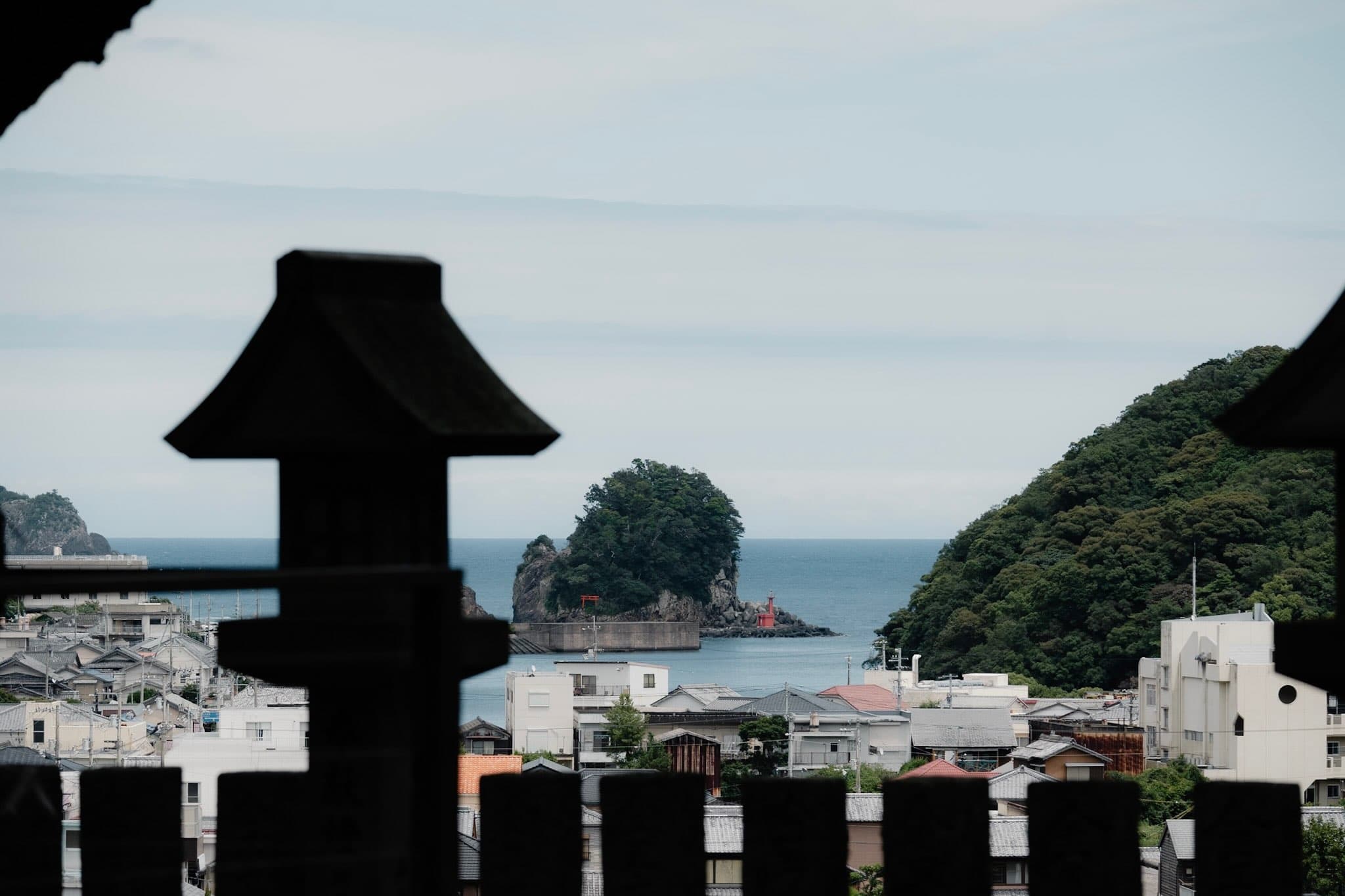 石灯籠と石柵の影の向こうに、瓦屋根の町並みと海、木に覆われた小島の岩場に立つ赤い灯台と鳥居が見える