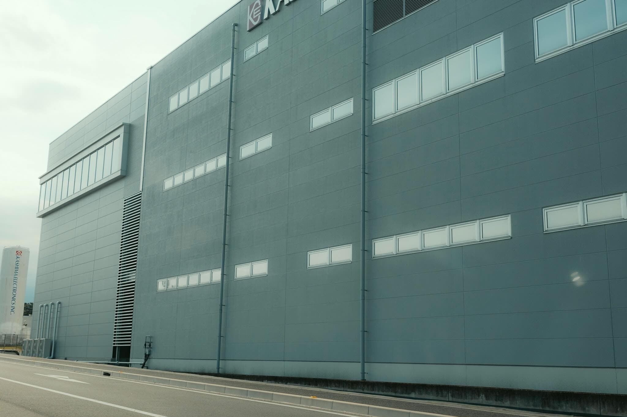 灰色の外壁に細長い窓が並ぶ工場建屋を道路から見上げた構図。左端に「KASHIMA ELECTRONICS INC.」のタンクが立つ