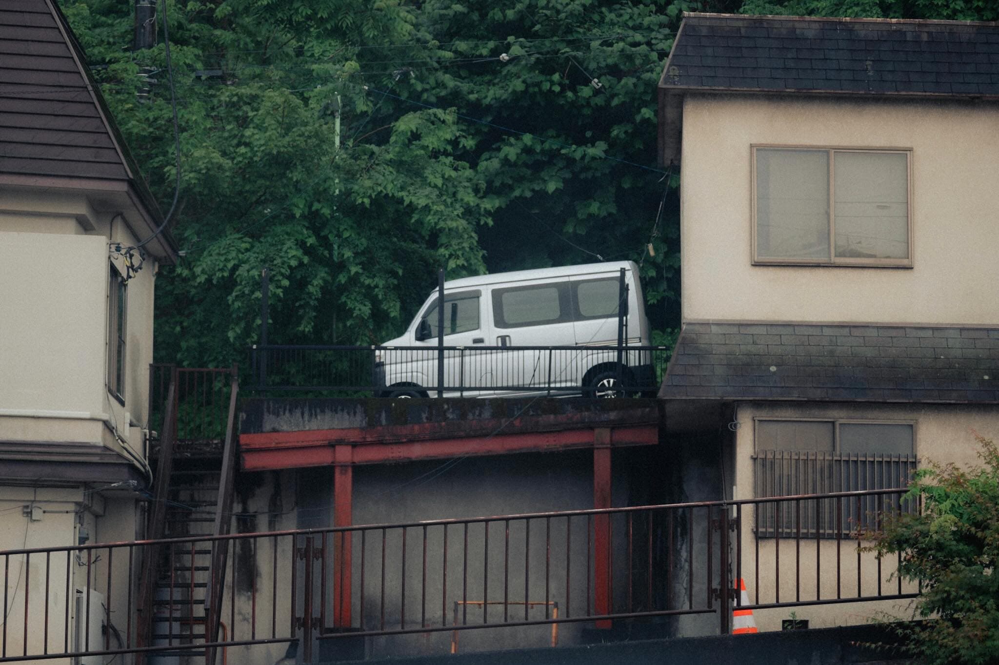 住宅の間の高架式駐車場に停められた白い軽バン。柵と赤い鉄骨の梁に支えられ、背後は緑の木立