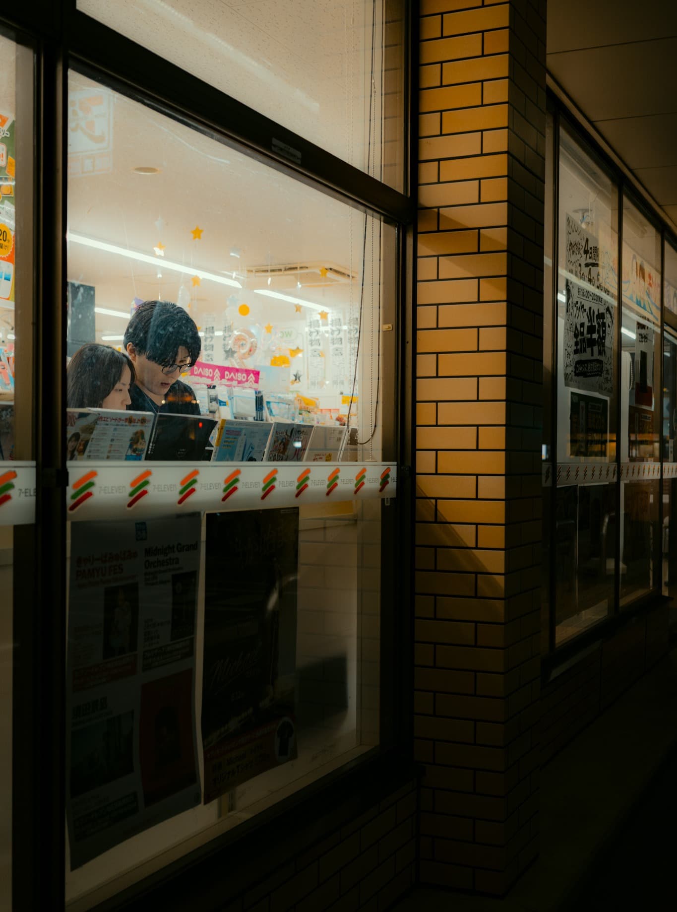 夜のコンビニの窓越しに、雑誌棚の前に立つ眼鏡の男性と女性。窓の下に7-ELEVENの帯と、ライブ告知のポスターが貼られている