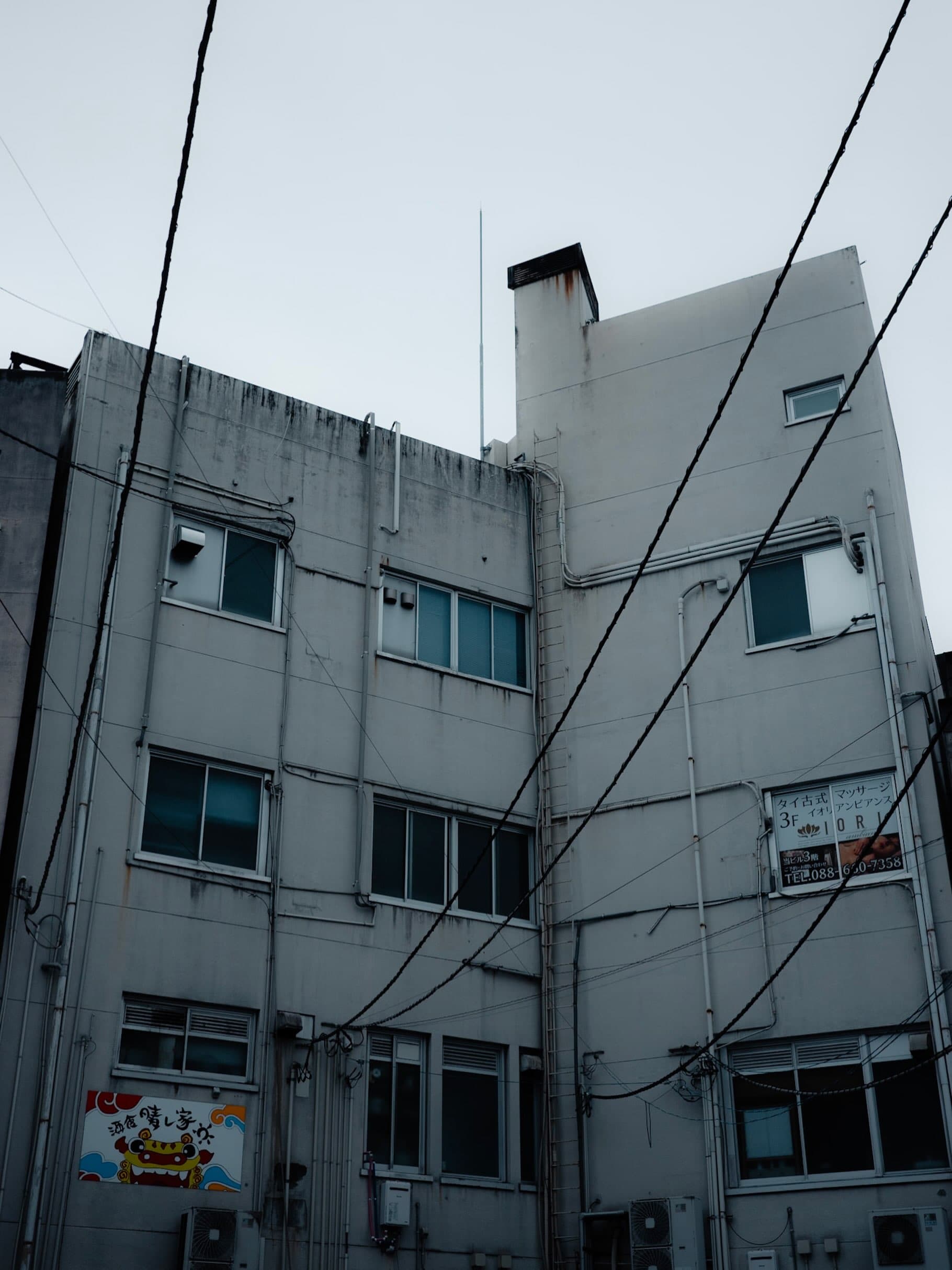 曇り空を背に見上げた四階建ての白いビル。何本もの電線が斜めに横切り、壁面に「タイ古式マッサージ」と「酒食 晴し家」の看板が掛かる